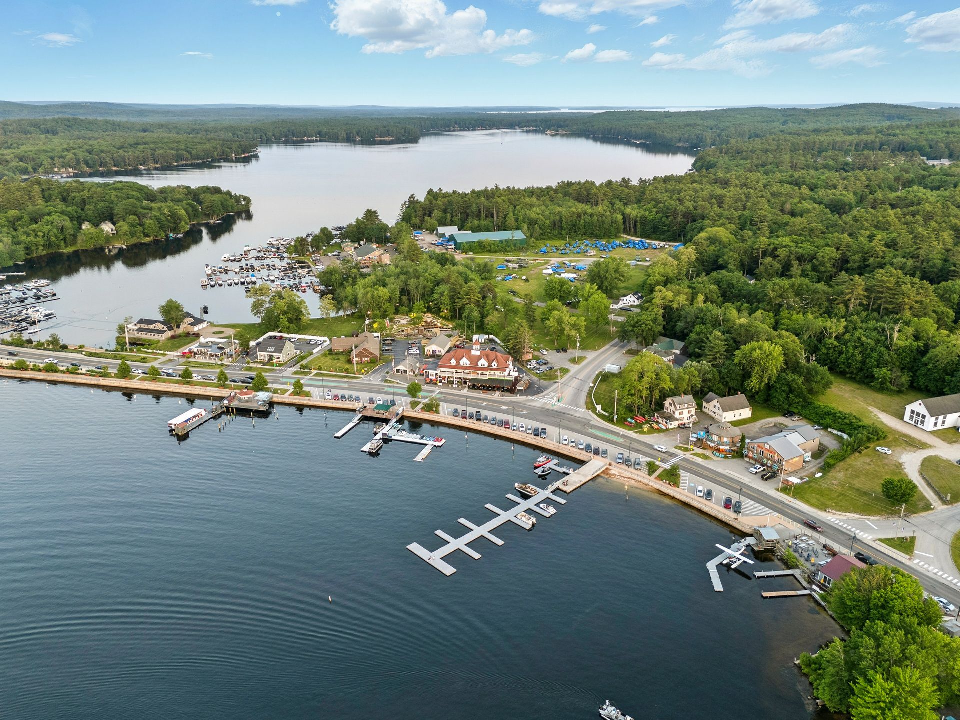 Aerial view of a lakeside town with docks, boats, buildings, and lush green trees under a blue sky.