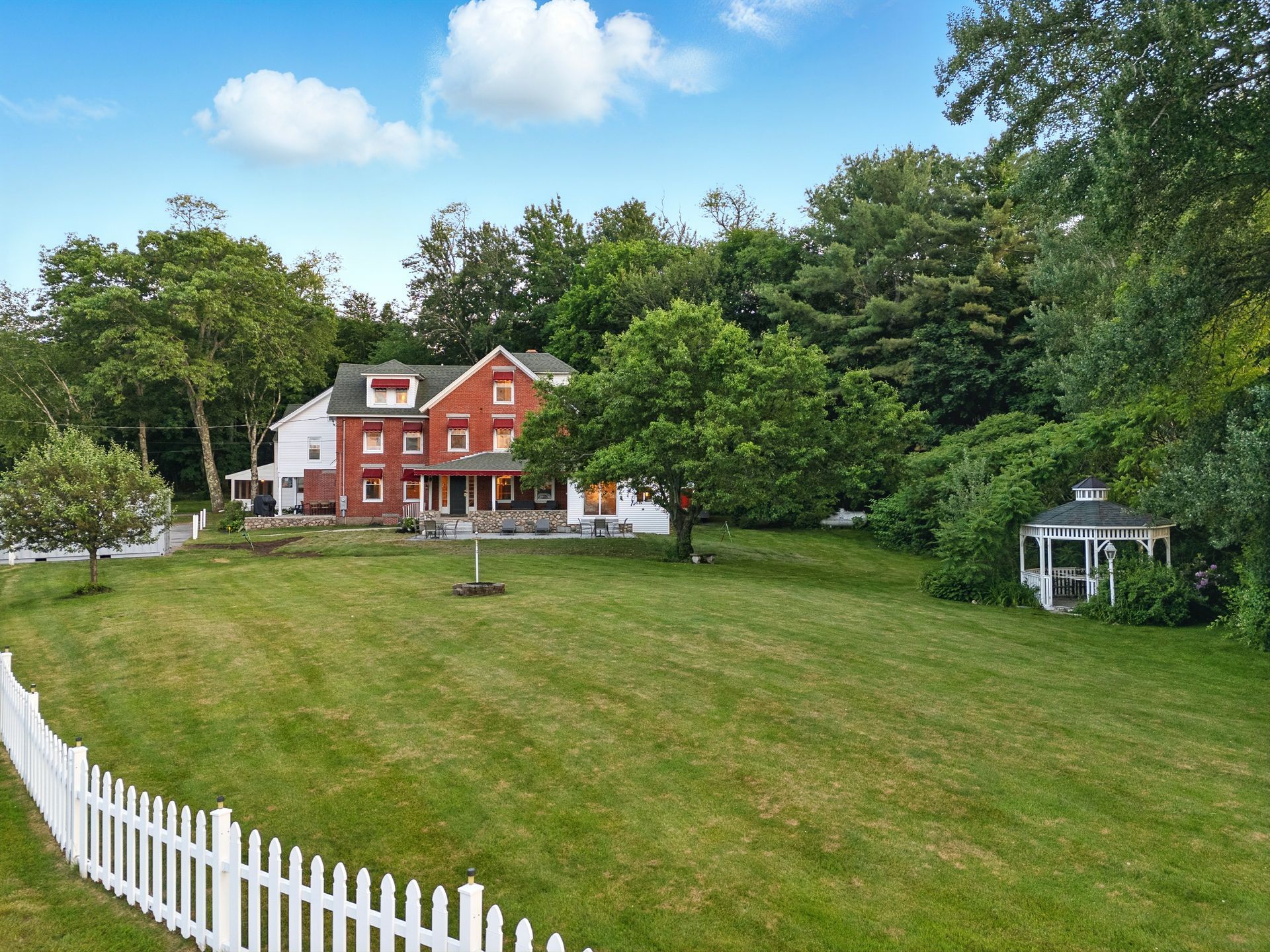Large red-brick and white house with a picket fence, gazebo, and green lawn. Trees surround the property.