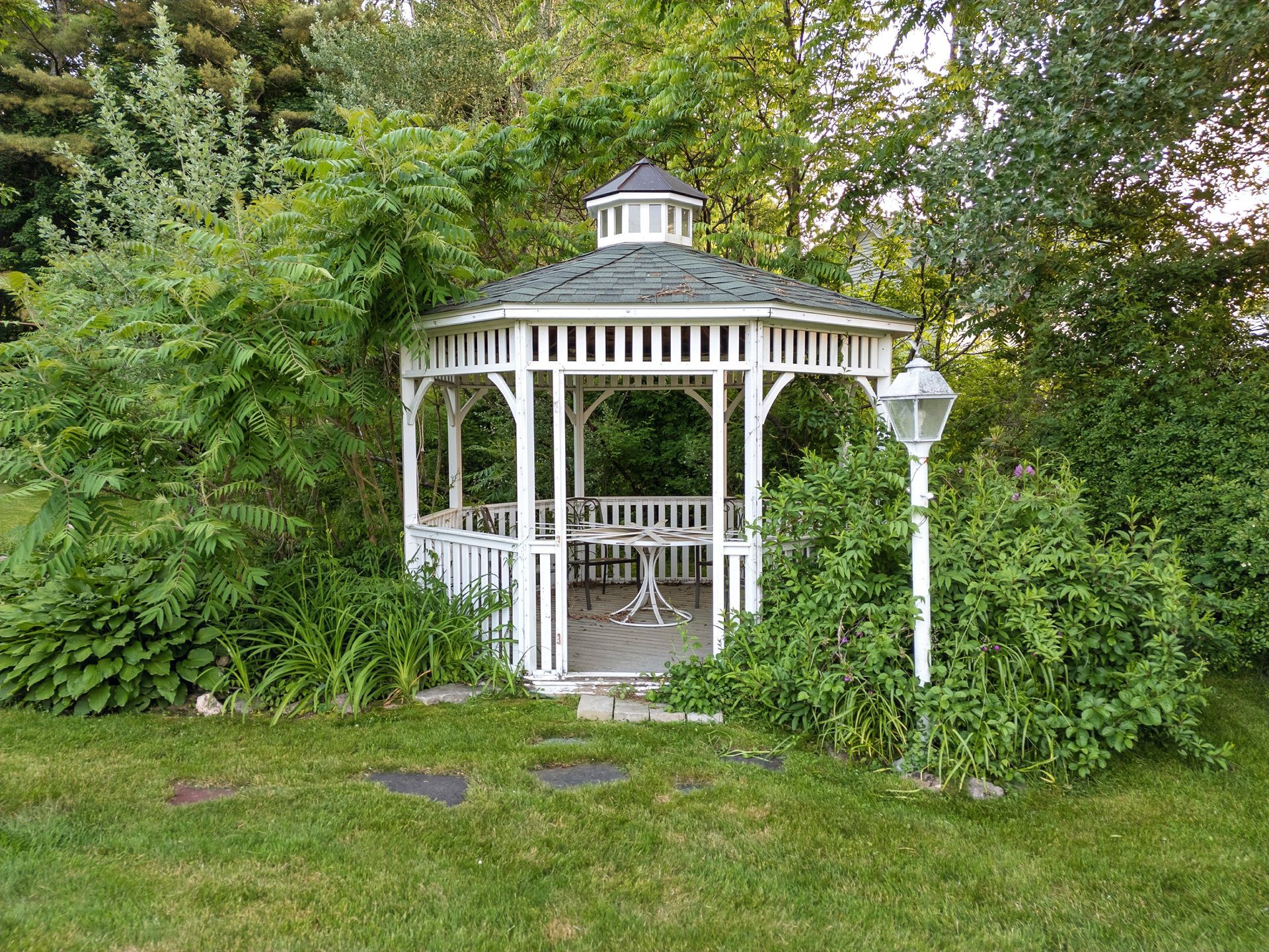 White gazebo surrounded by greenery, on a grassy lawn with stepping stones.