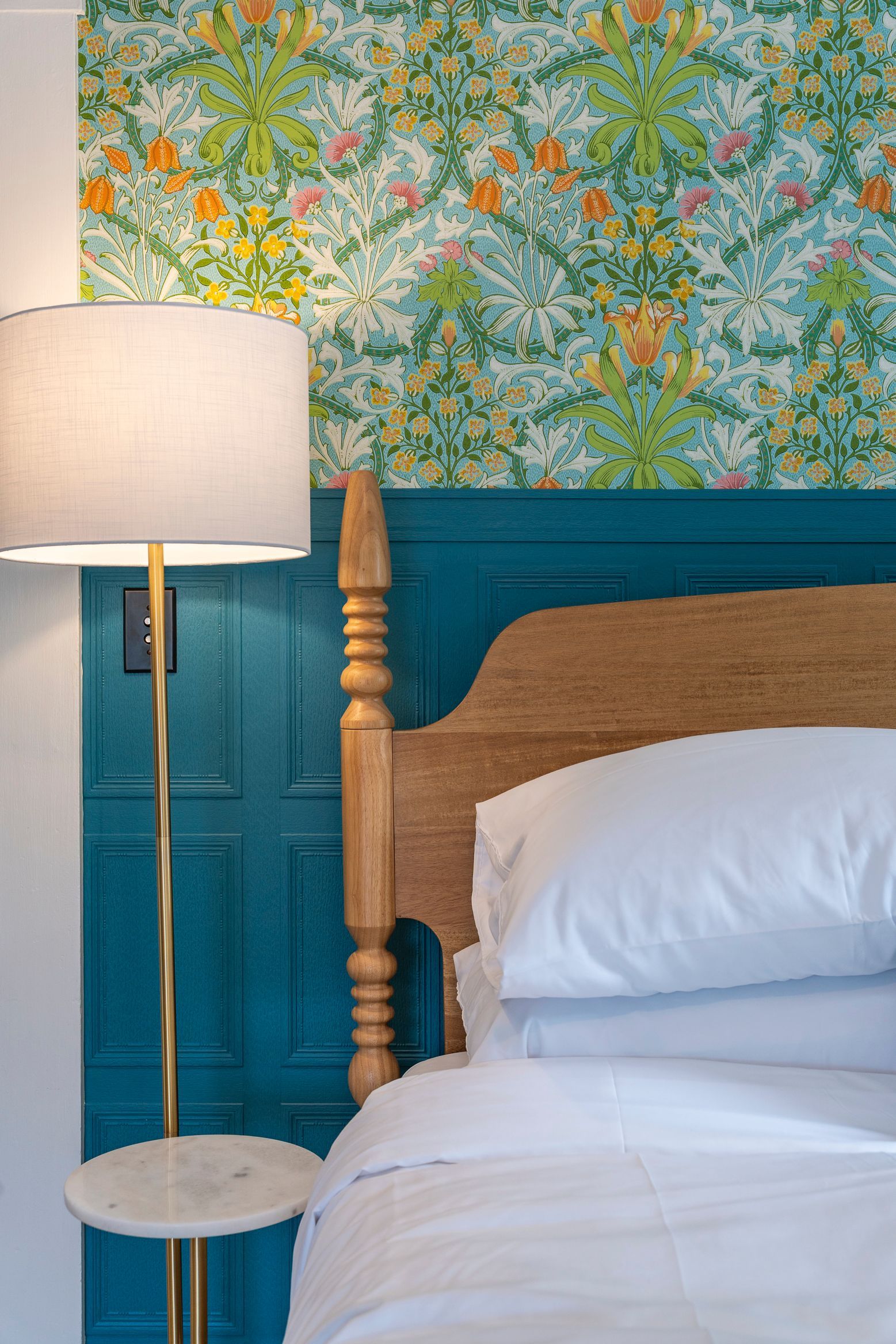 Bedroom with a bed, a teal headboard, floral wallpaper, and a gold lamp with a white shade.