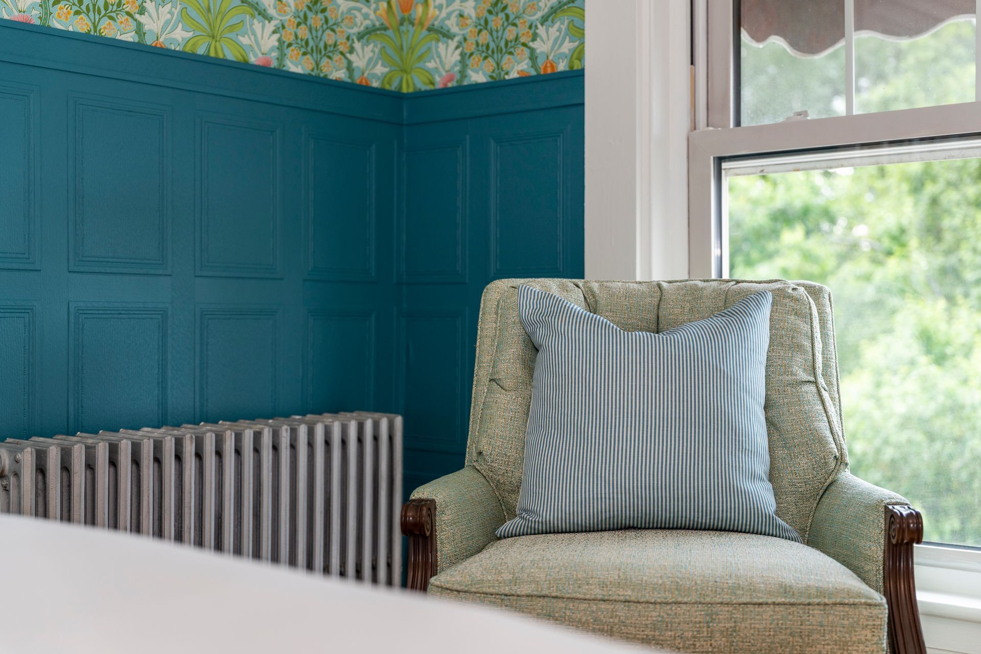 Teal paneled wall, floral wallpaper corner, armchair with blue striped pillow, radiator, and window with greenery.