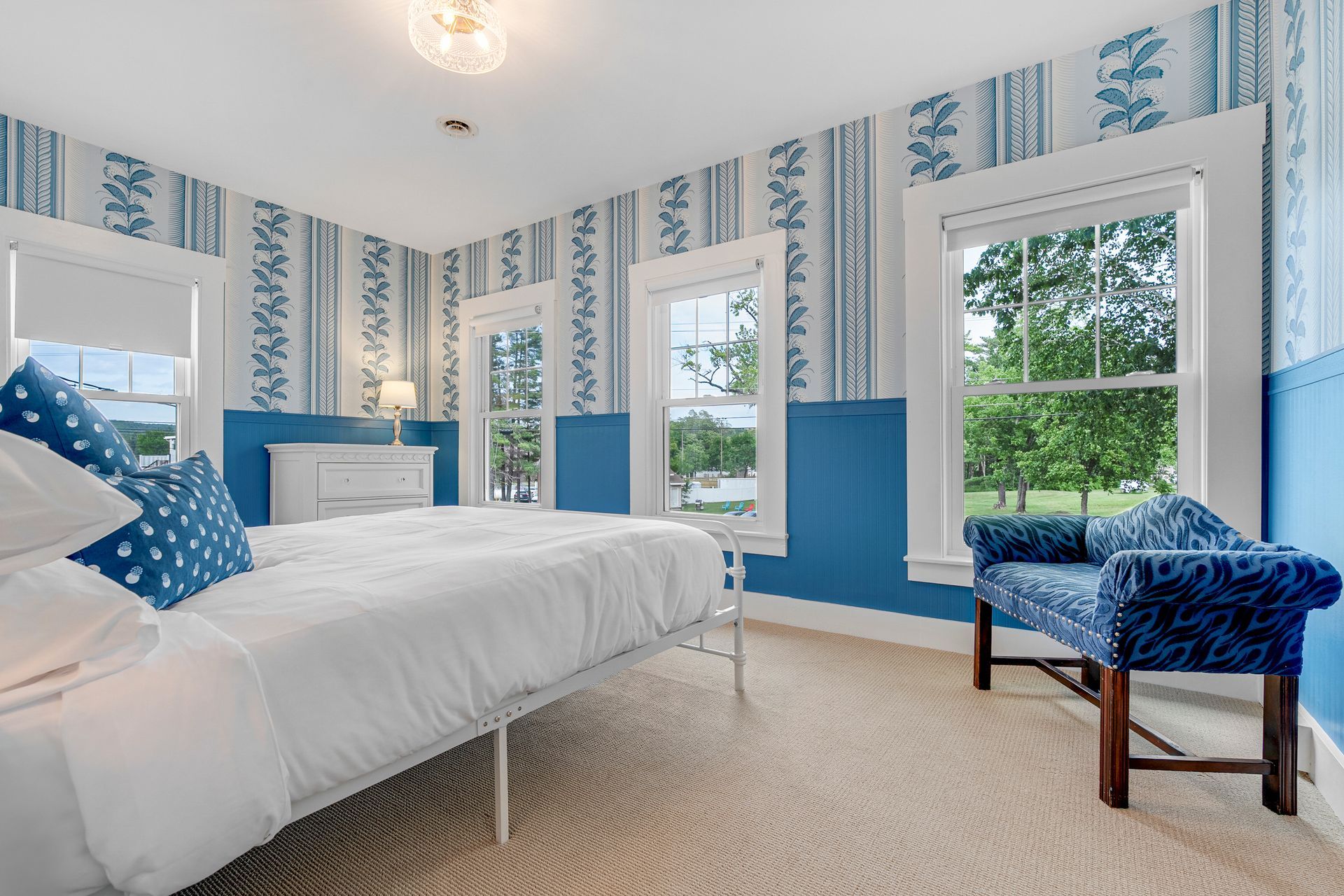 Bedroom with blue and white patterned wallpaper, a white bed, and a blue patterned chair near a window.