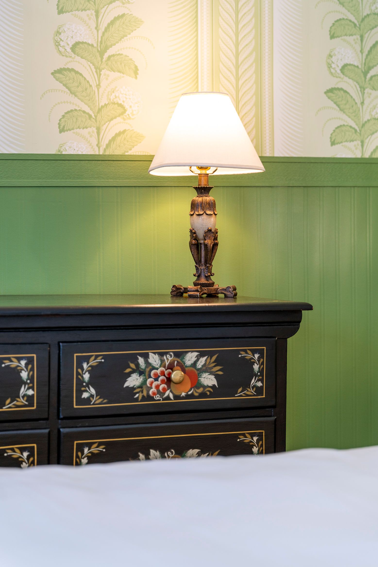 Black nightstand with floral accents, topped by a lamp with a lampshade, against green and patterned wallpaper.