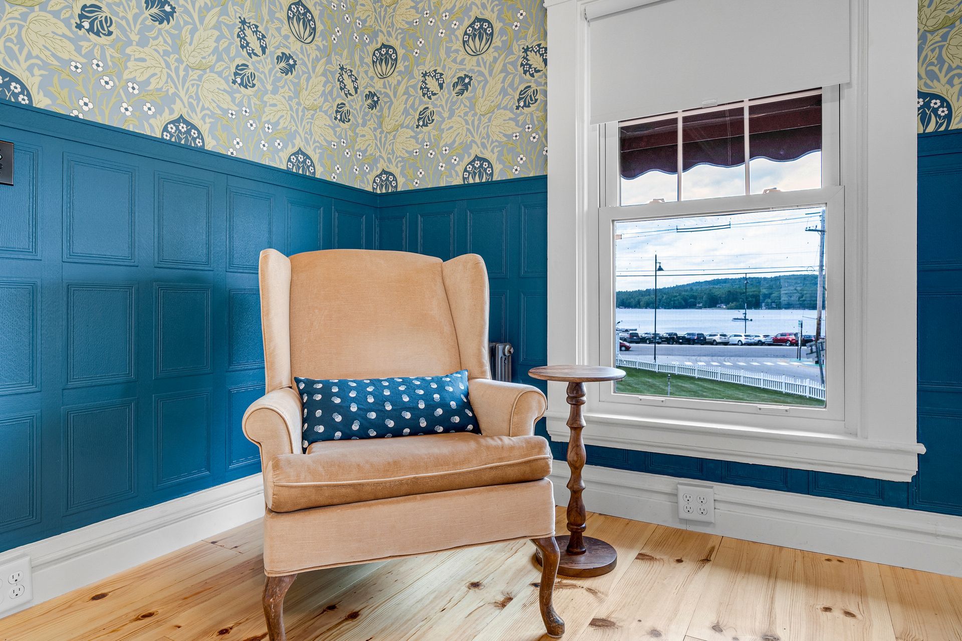 Cozy room with blue paneling, patterned wallpaper, a tan armchair, small table, and a window with a waterfront view.