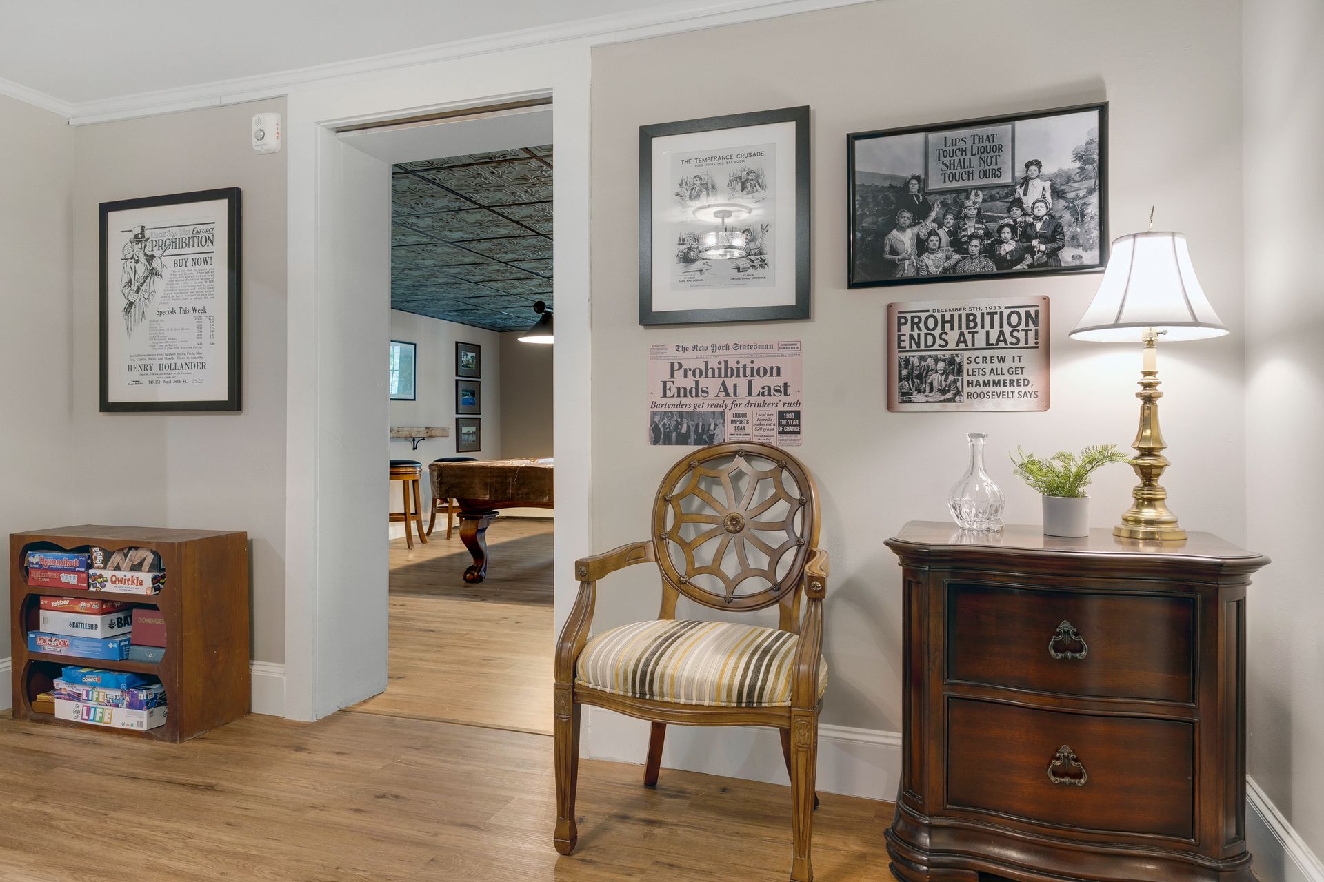Room with framed art, wooden furniture, and doorway to a pool table room. Light wood floors, neutral walls.