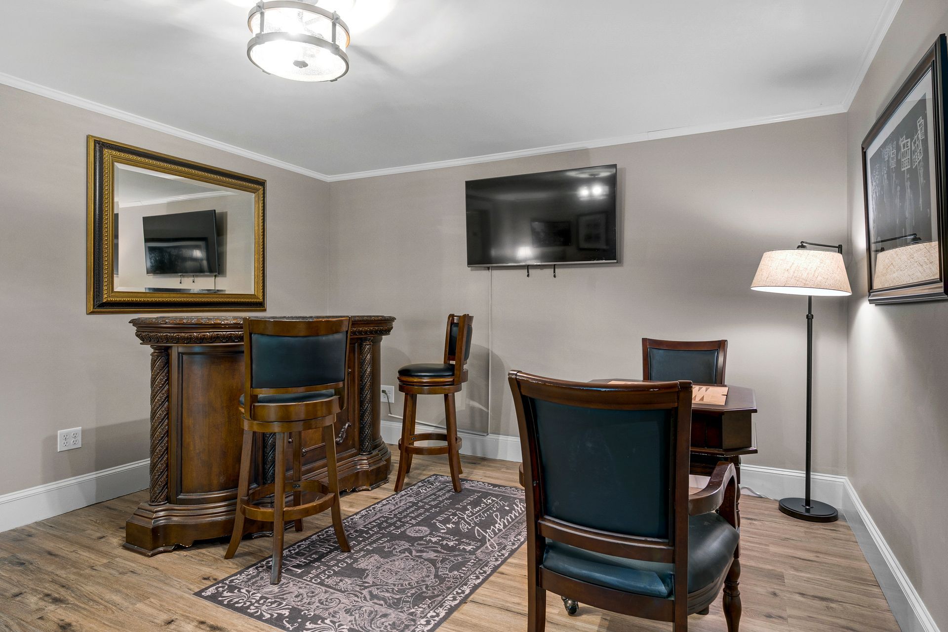 Basement entertainment area with a bar, seating, TV, and lamp. The walls are gray, and the floor is wood.