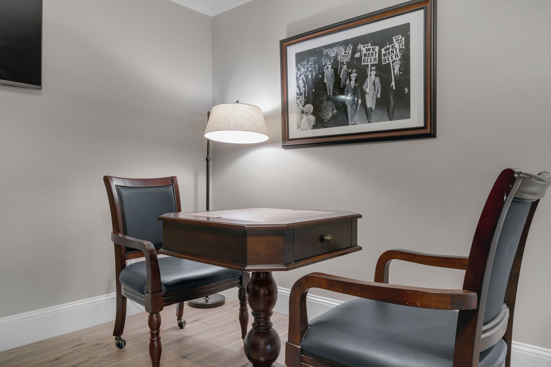 Two chairs face a small table beneath a photo and a floor lamp in a room with light gray walls.