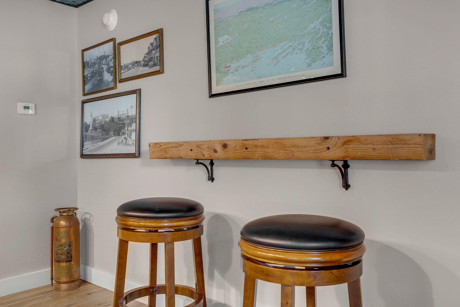 Two stools sit beneath a wooden shelf with framed pictures on a gray wall.