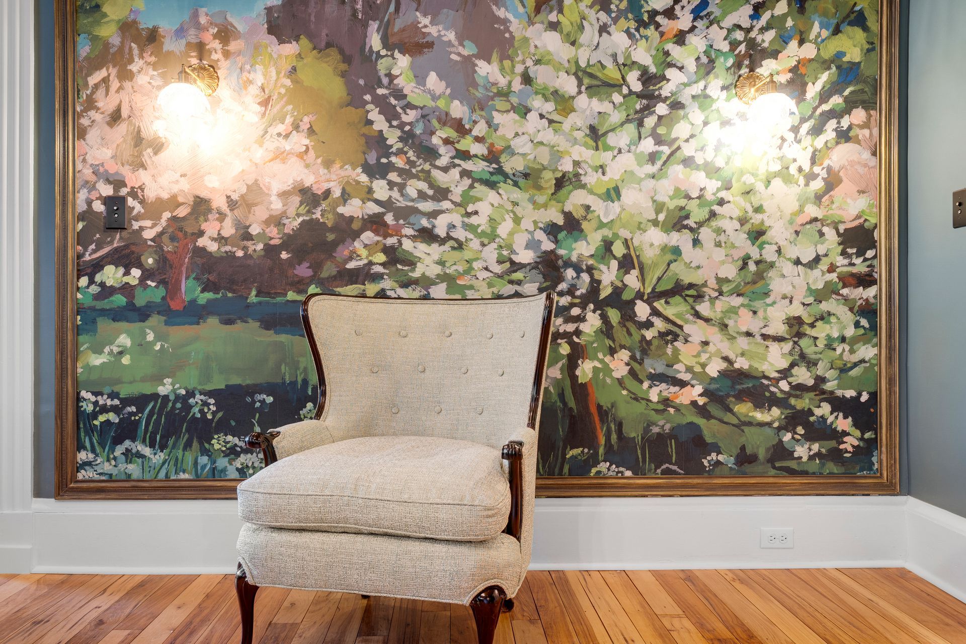 Chair in front of large floral painting, hardwood floor, two sconces on wall.