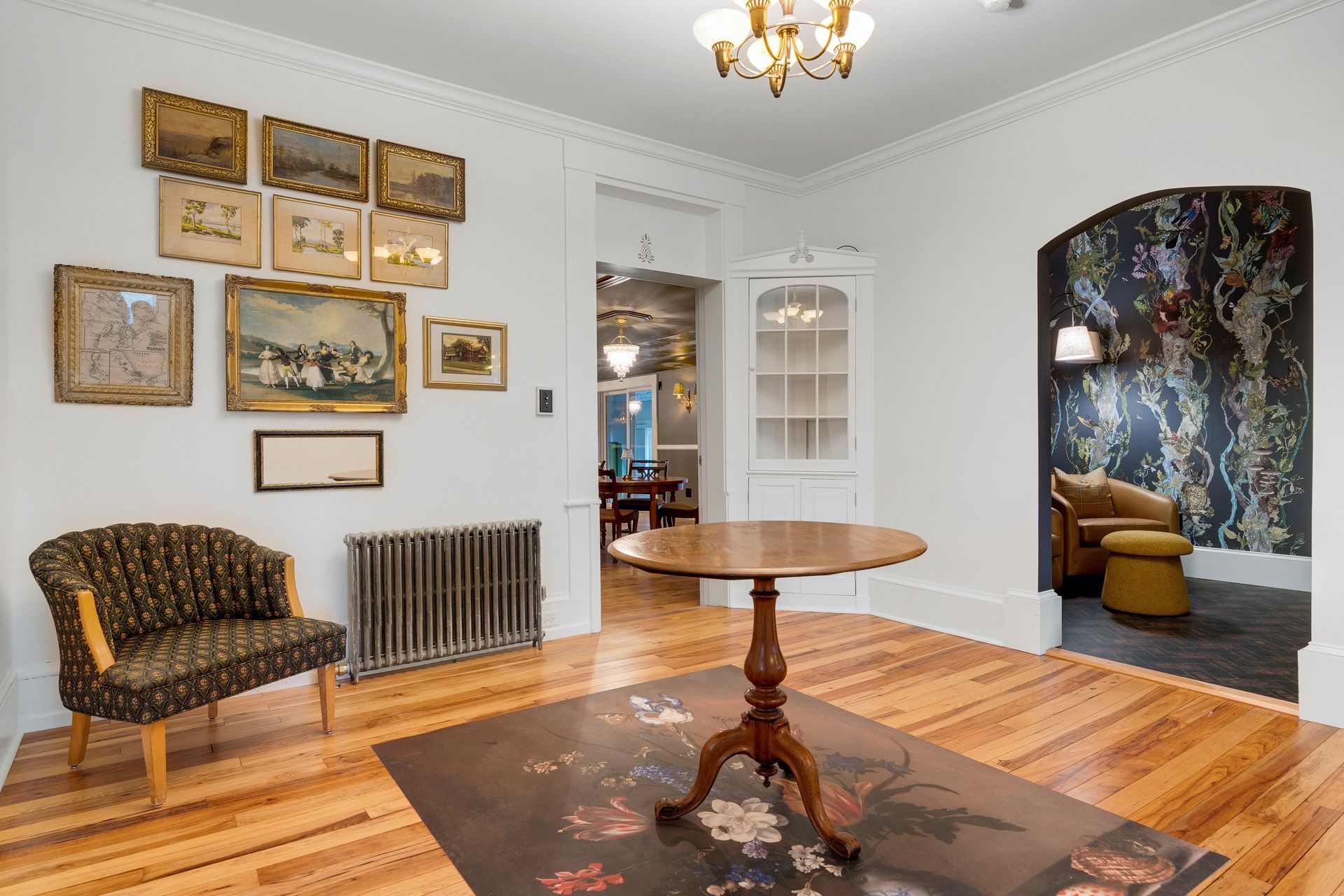 A room with antique furniture, wooden floors, and floral wallpaper. Artwork adorns the walls.