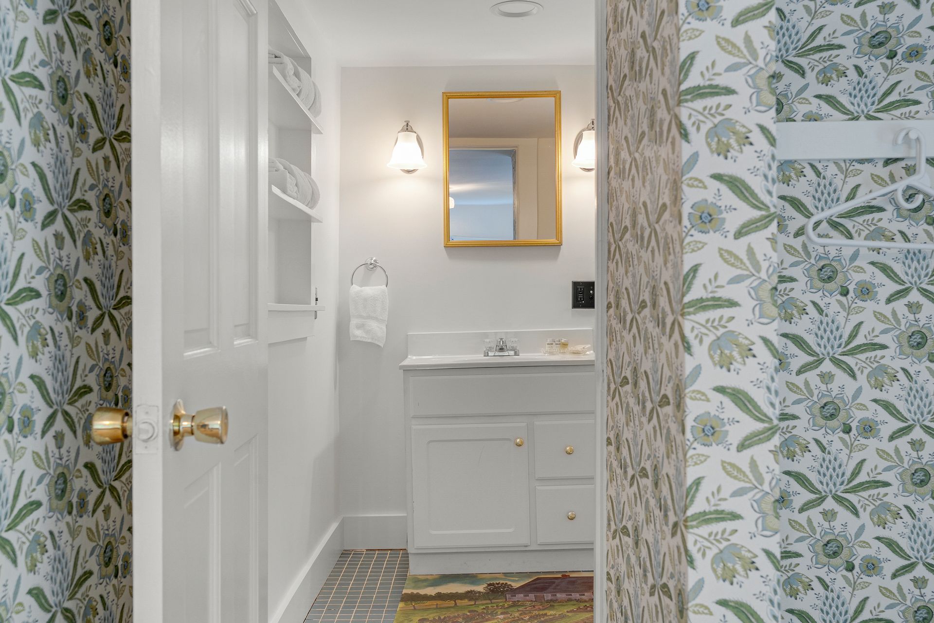 Small white bathroom with floral wallpaper, vanity, gold mirror, and open doorway.