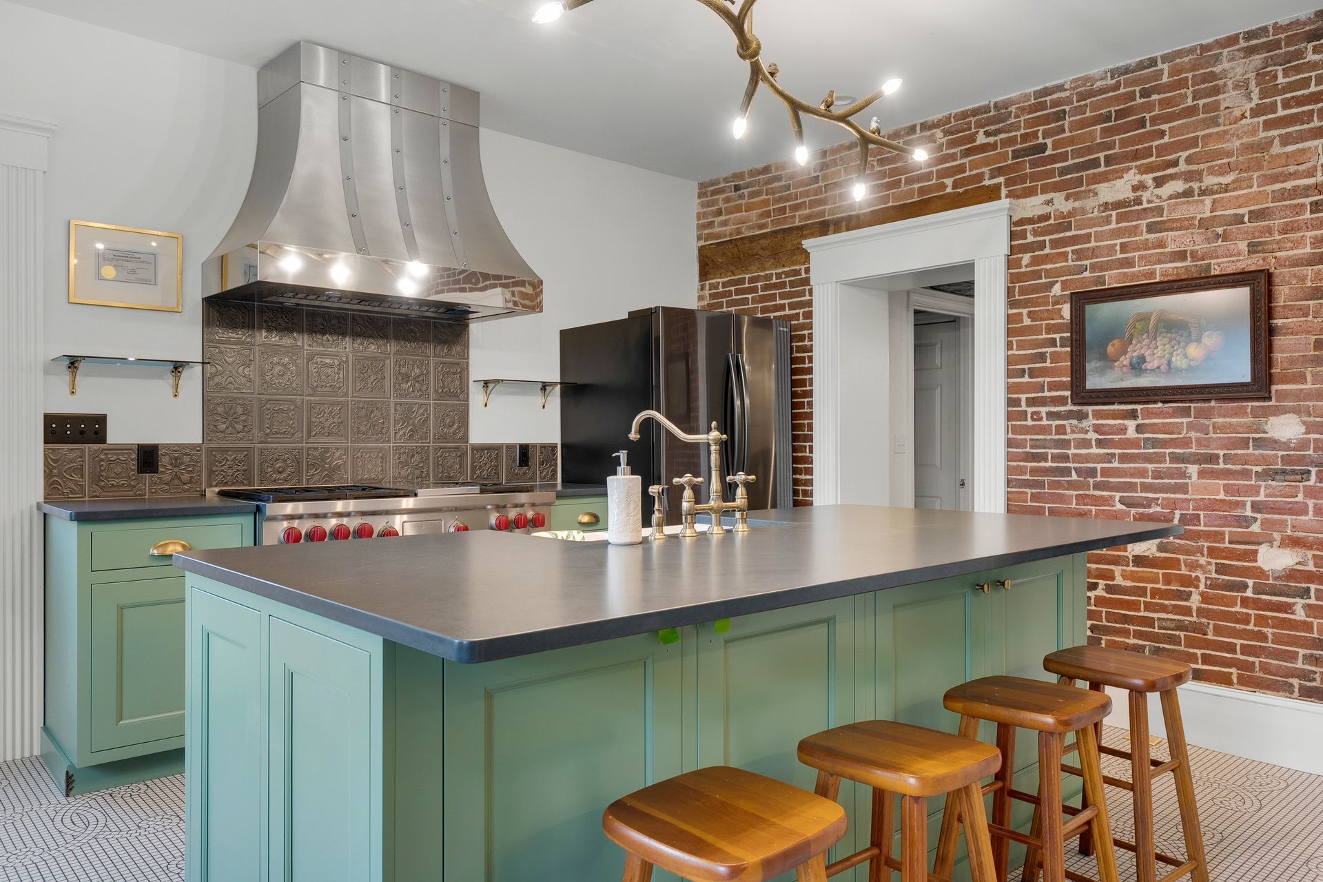Kitchen with green island, stainless hood, brick wall, and wooden stools.