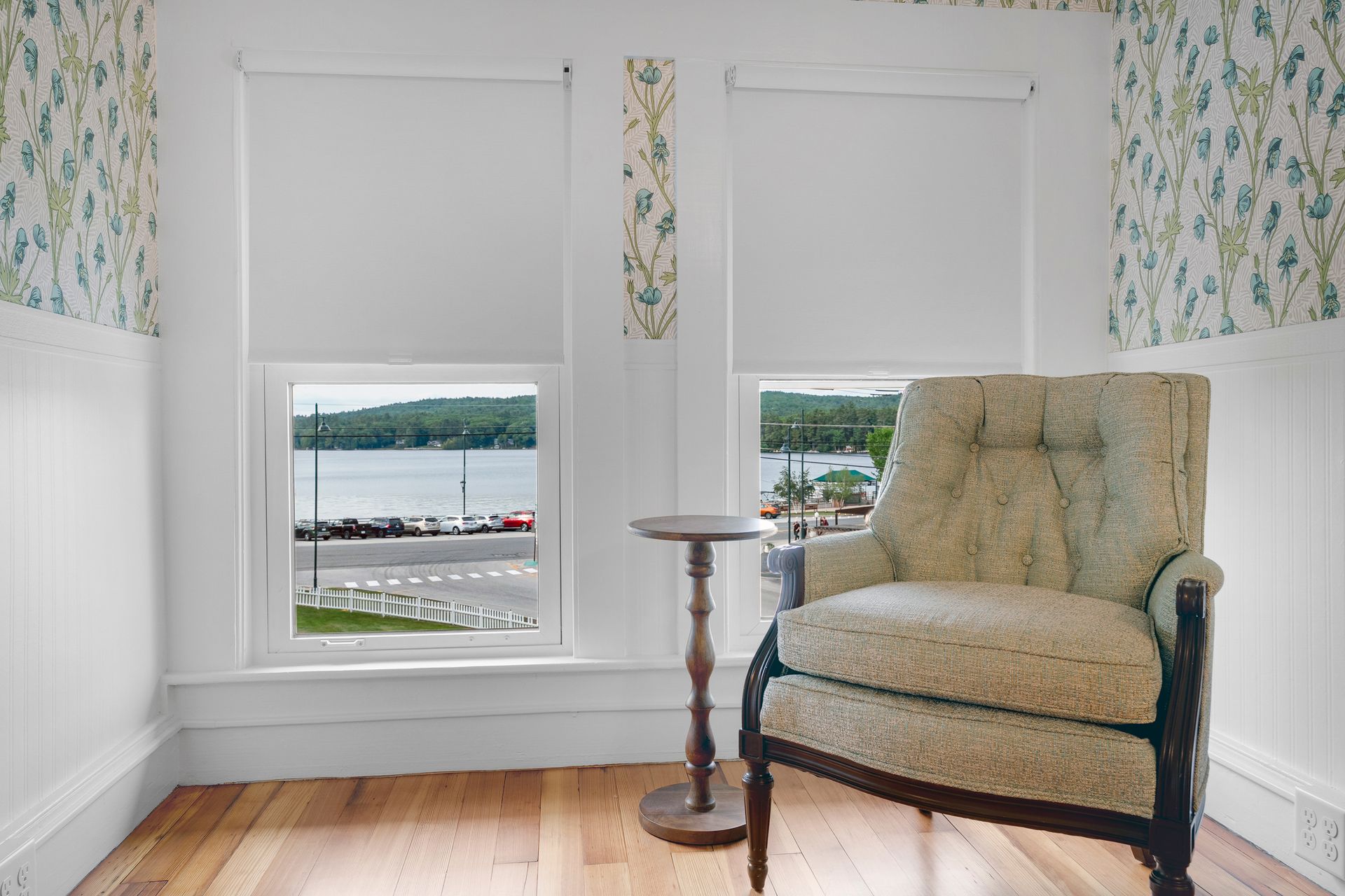 Cozy room with two windows overlooking a lake. A patterned armchair sits beside a small table. Floral wallpaper.