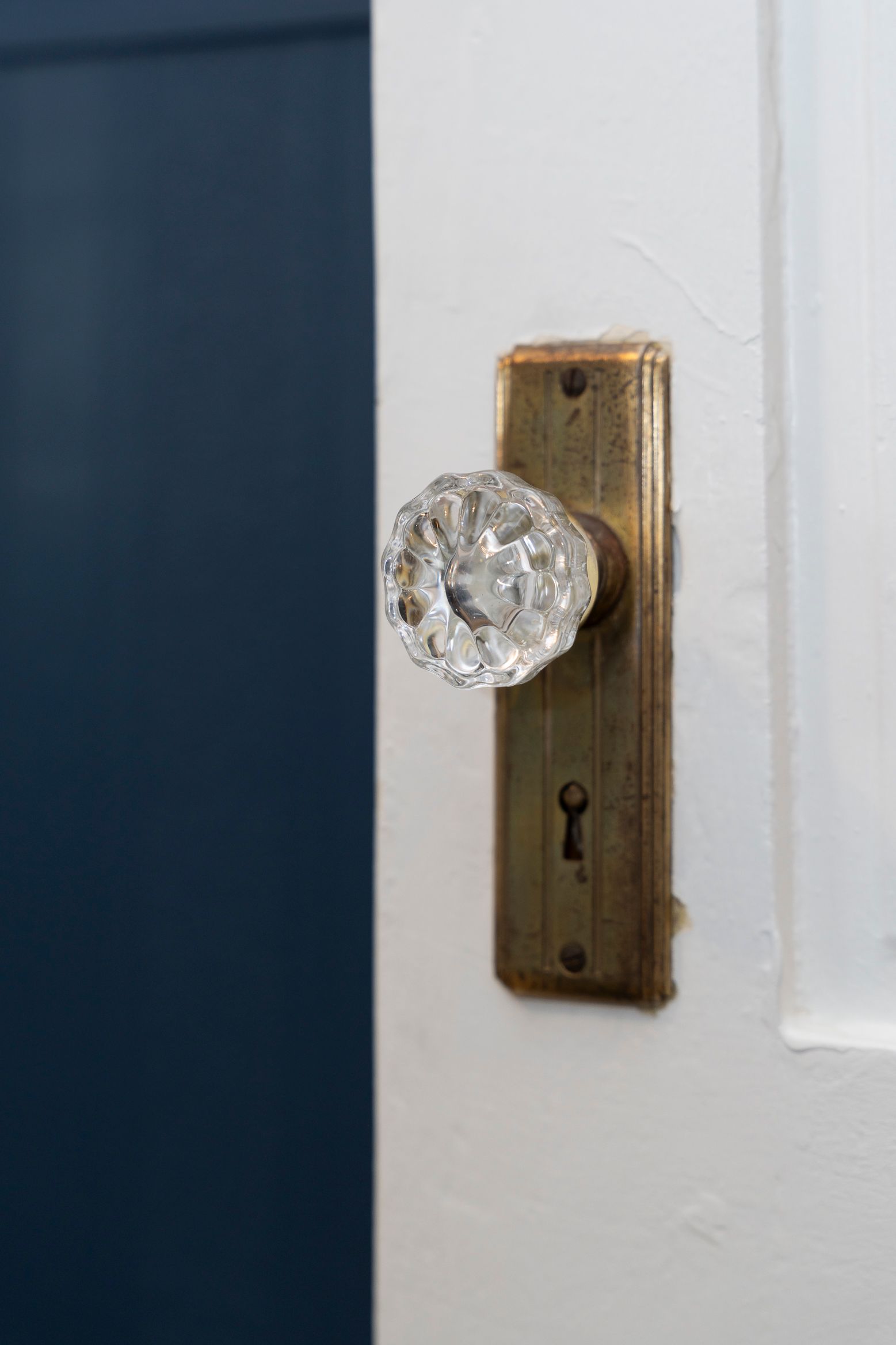 Crystal doorknob on aged brass plate set on white door, opening to a navy blue wall.