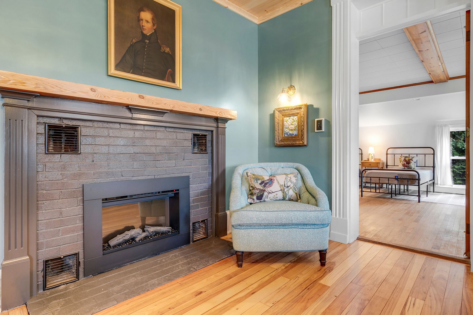 Fireplace with portrait and chair in front; doorway to bedroom with bed.
