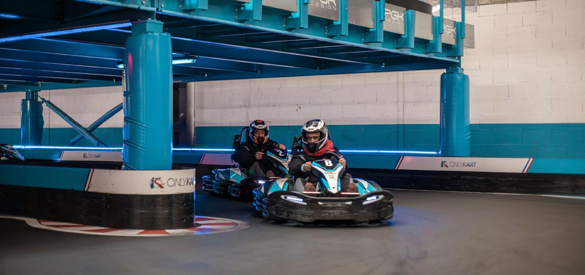 Two people in racing karts on a track under a blue structure. They wear helmets and appear to be racing.
