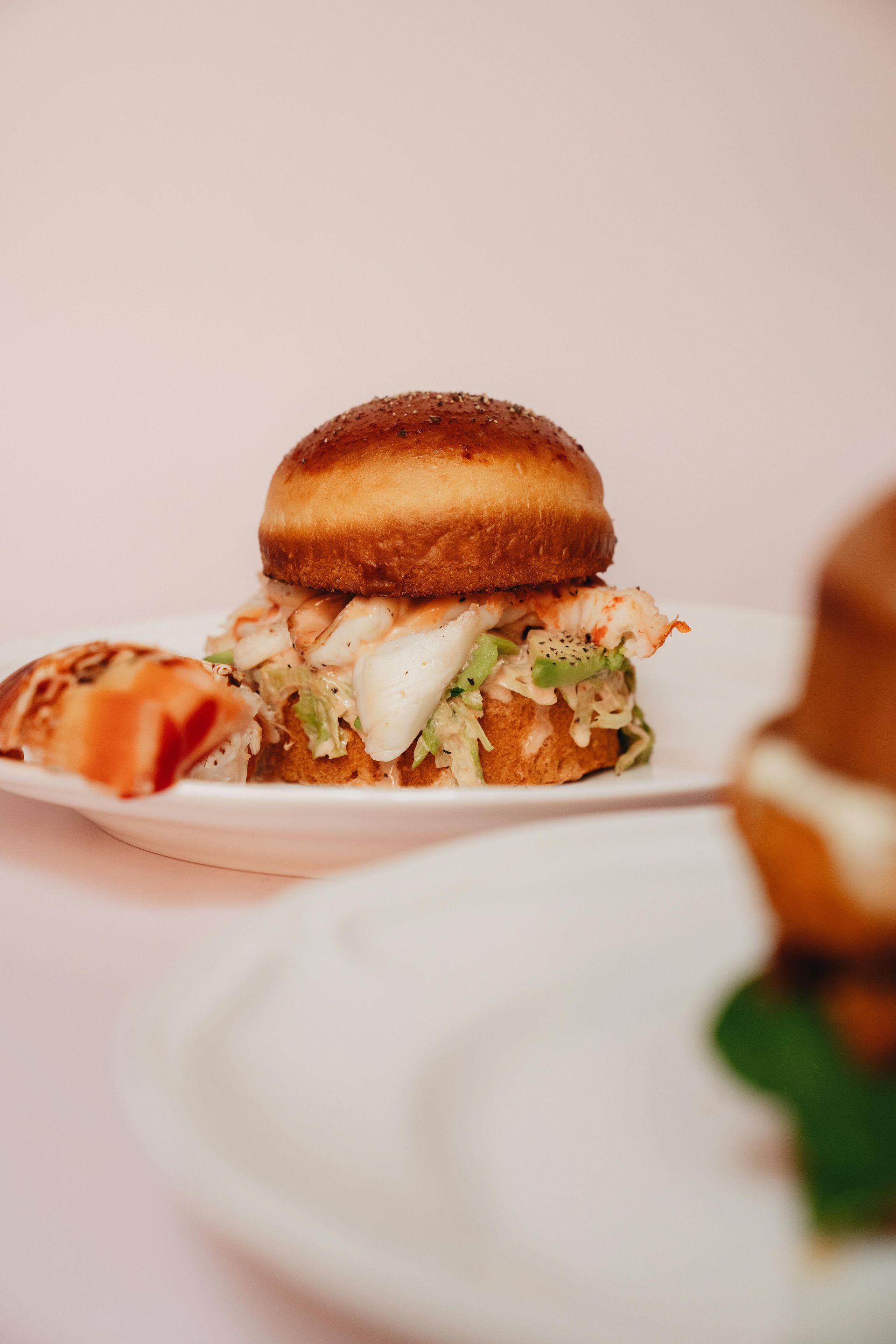Lobster slider on a white plate with slaw and a golden bun, lit with soft pink.