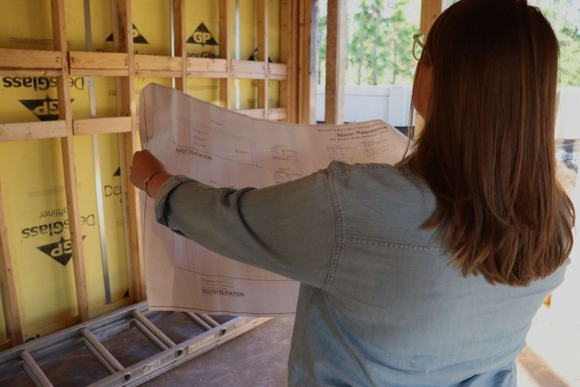 a woman holding and analizing a blueprint with her hands. In the backgroung there is a house under construction with the structural made out of wood.