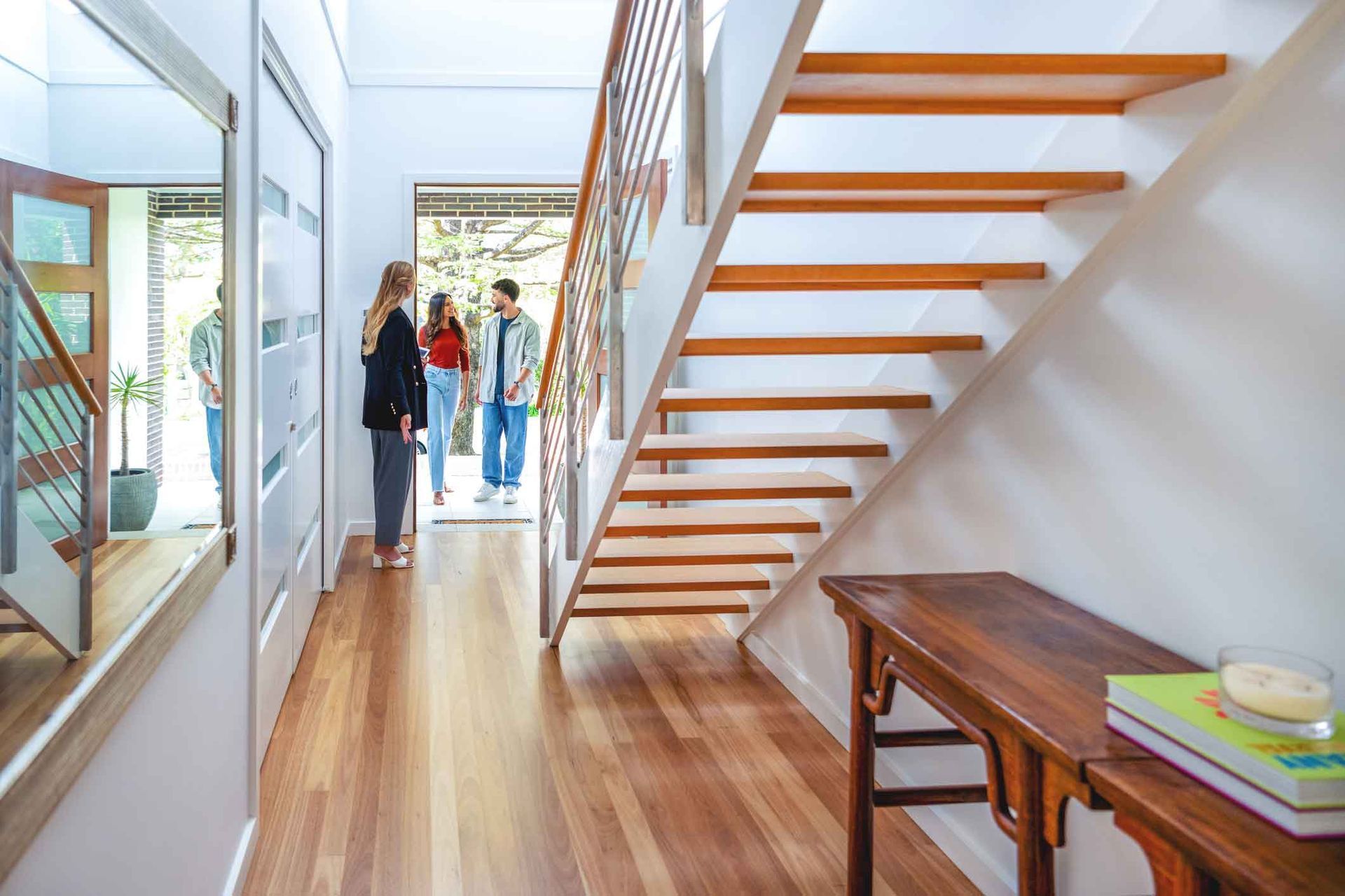 A couple of people are standing in a hallway next to a wooden staircase. A couple of people are standing in a hallway next to a wooden staircase.