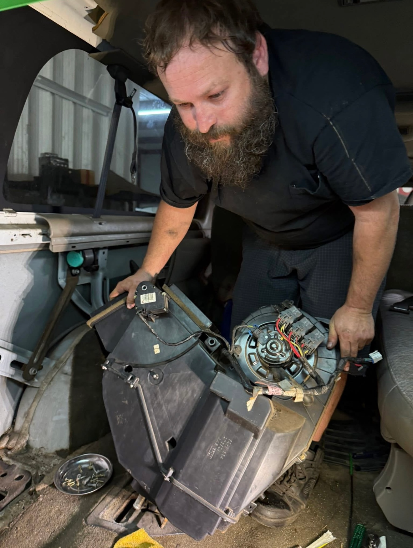 A bearded mechanic holding a car blower motor assembly inside a vehicle cabin. | Bucks Auto Garage & Tire