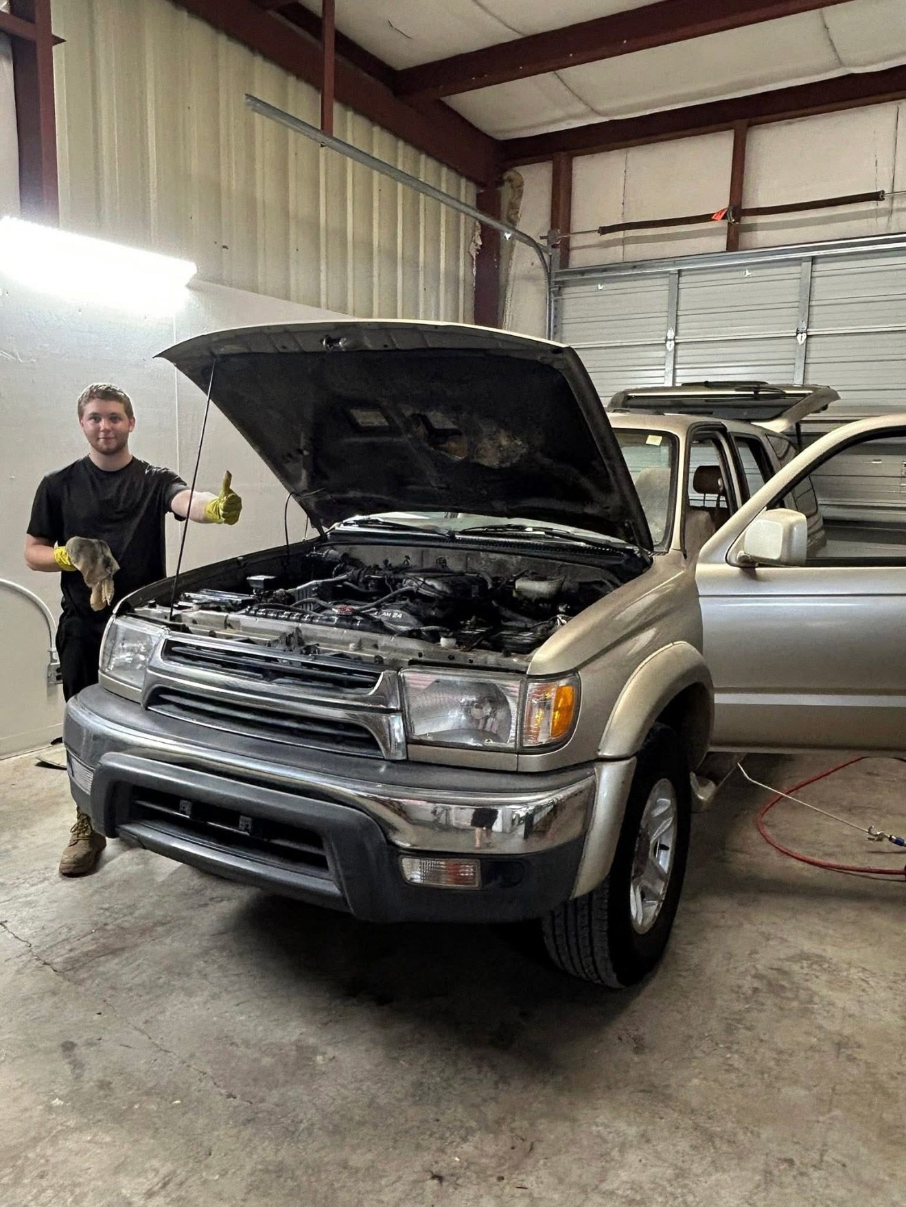 A person in a garage stands with a thumbs up next to a silver SUV with its hood open for engine work. | Bucks Auto Garage & Tire