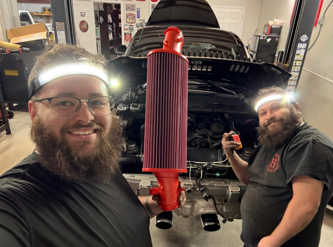 Two people wearing headlamps smile in a workshop, holding a large, red, cylindrical automotive air filter. | Bucks Auto Garage & Tire