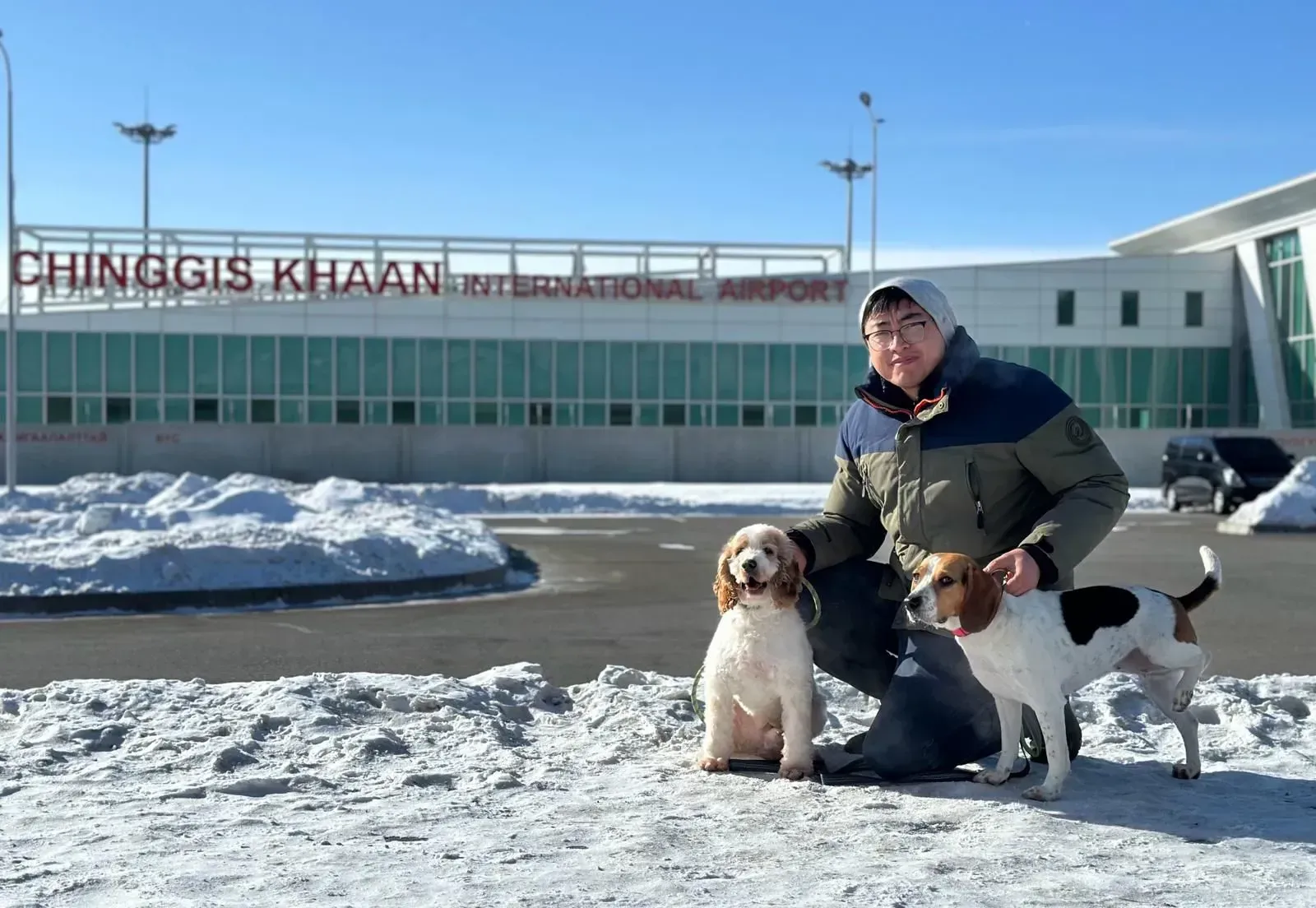 Man posing with two dogs outside Chinggis Khaan International Airport on a snowy day