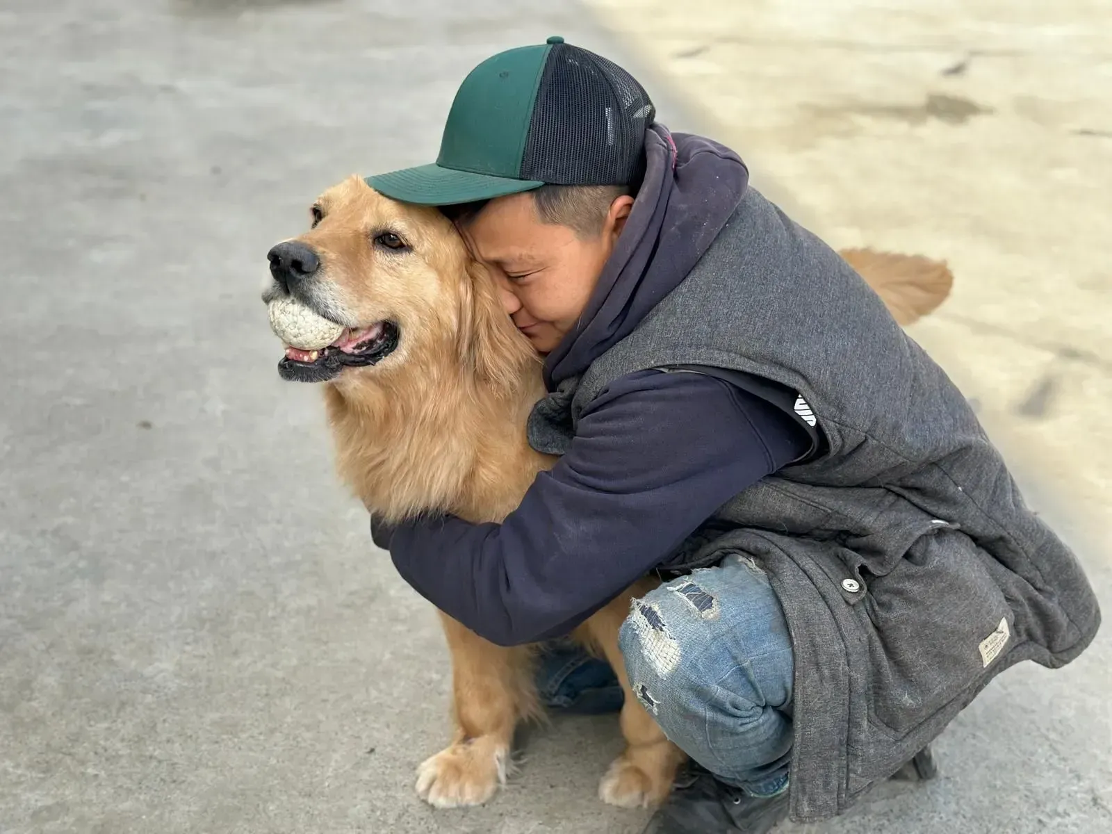 A child in a green cap hugs a golden dog outdoors on pavement.