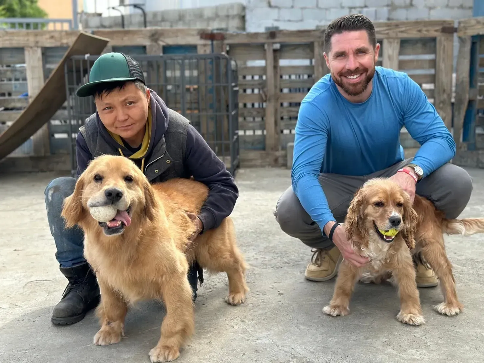 Two people kneel with two golden retrievers in a fenced outdoor area, all facing the camera and smiling.