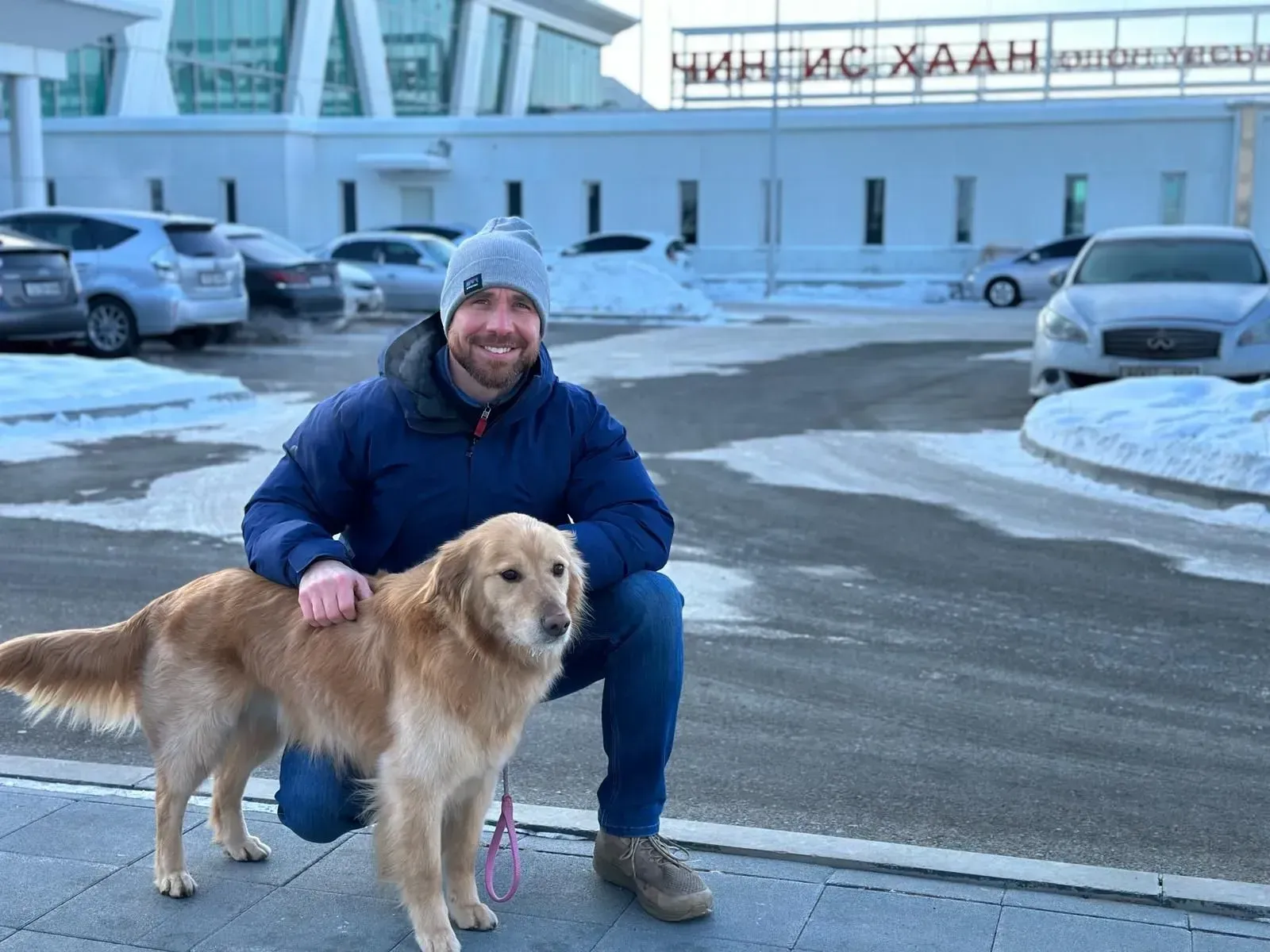 Man kneeling with a golden retriever on a snowy street in front of a building
