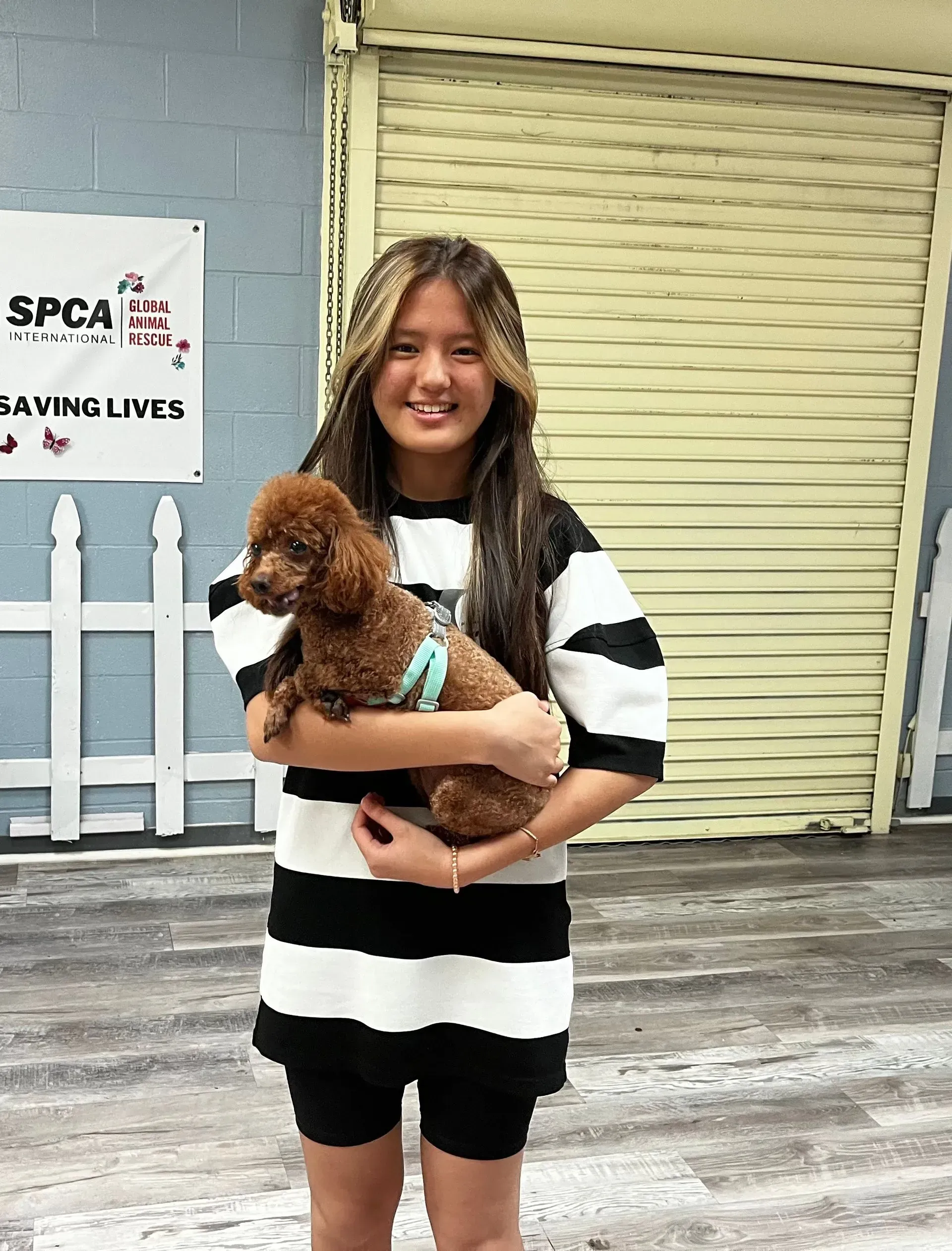 Young woman holding a small brown dog in front of a storefront with a white fence and closed shutter.