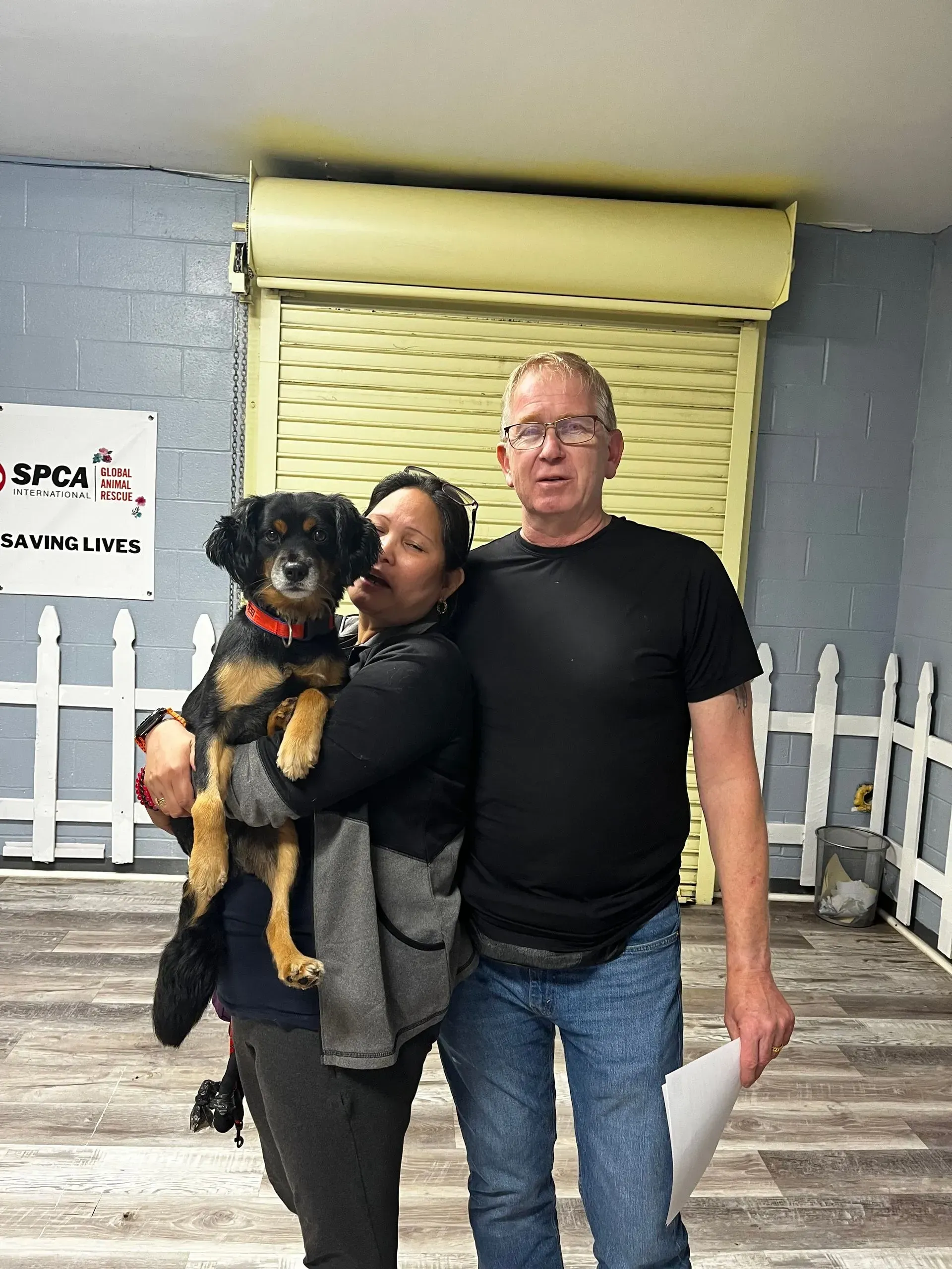 Two people posing indoors with a black dog in front of a yellow shutter and white fence backdrop