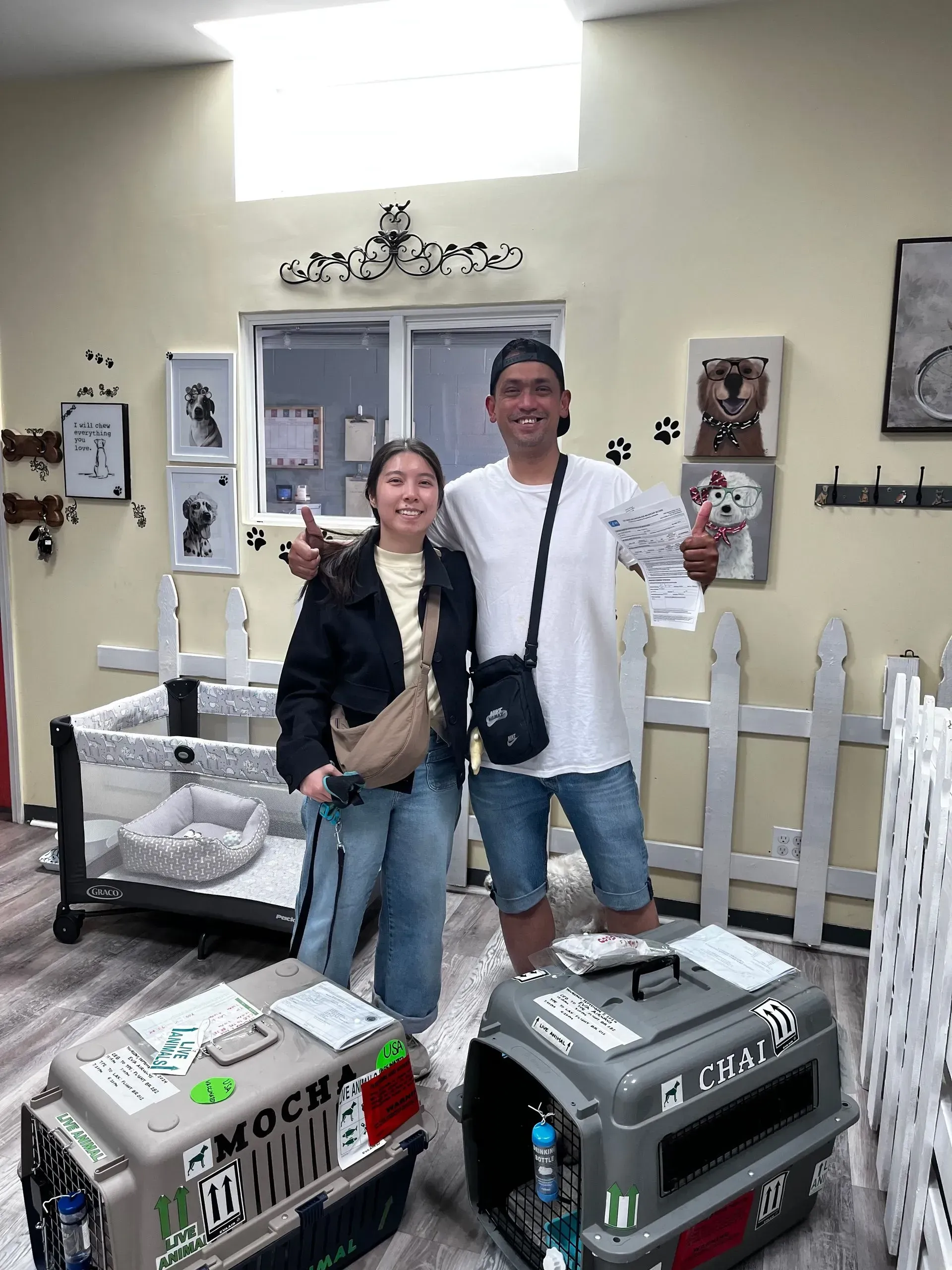 Two people smiling indoors beside two pet carriers and a white picket fence display.
