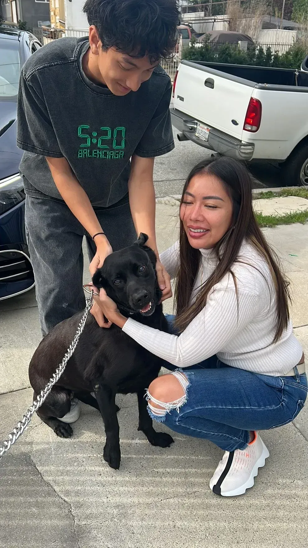 Two people crouch beside a black dog on a sidewalk, smiling near a parked white pickup truck.