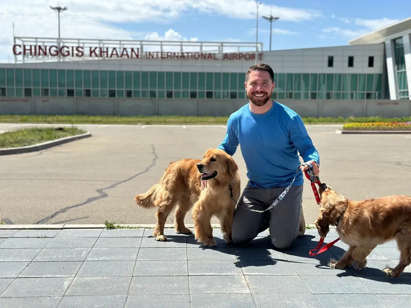 Man kneeling with two golden dogs and a brown dog outside Chinggis Khaan International Airport