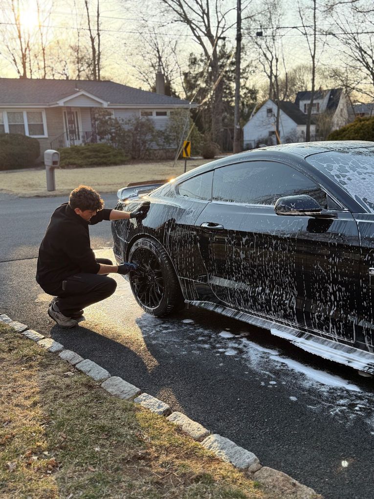 A man is kneeling down to wash a black car on the side of the road.