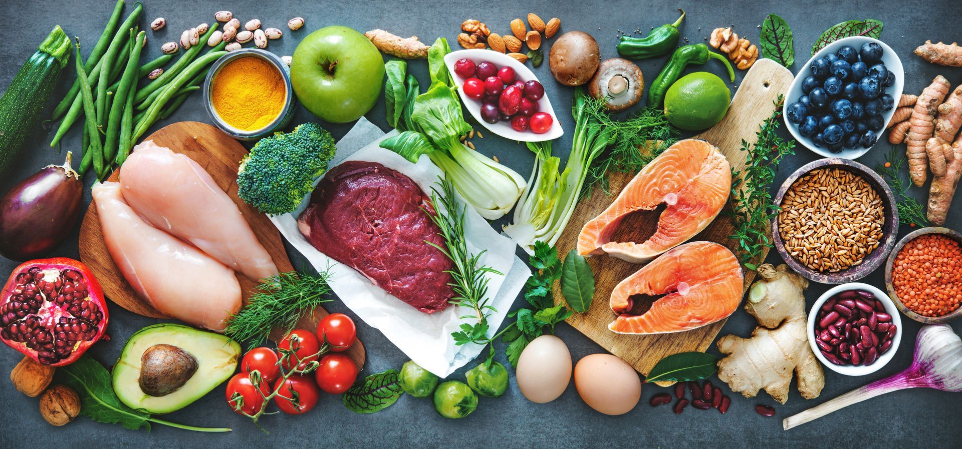 Assortment of fresh produce: meats, fruits, vegetables, and grains, arranged on a gray surface, emphasizing healthy eating.