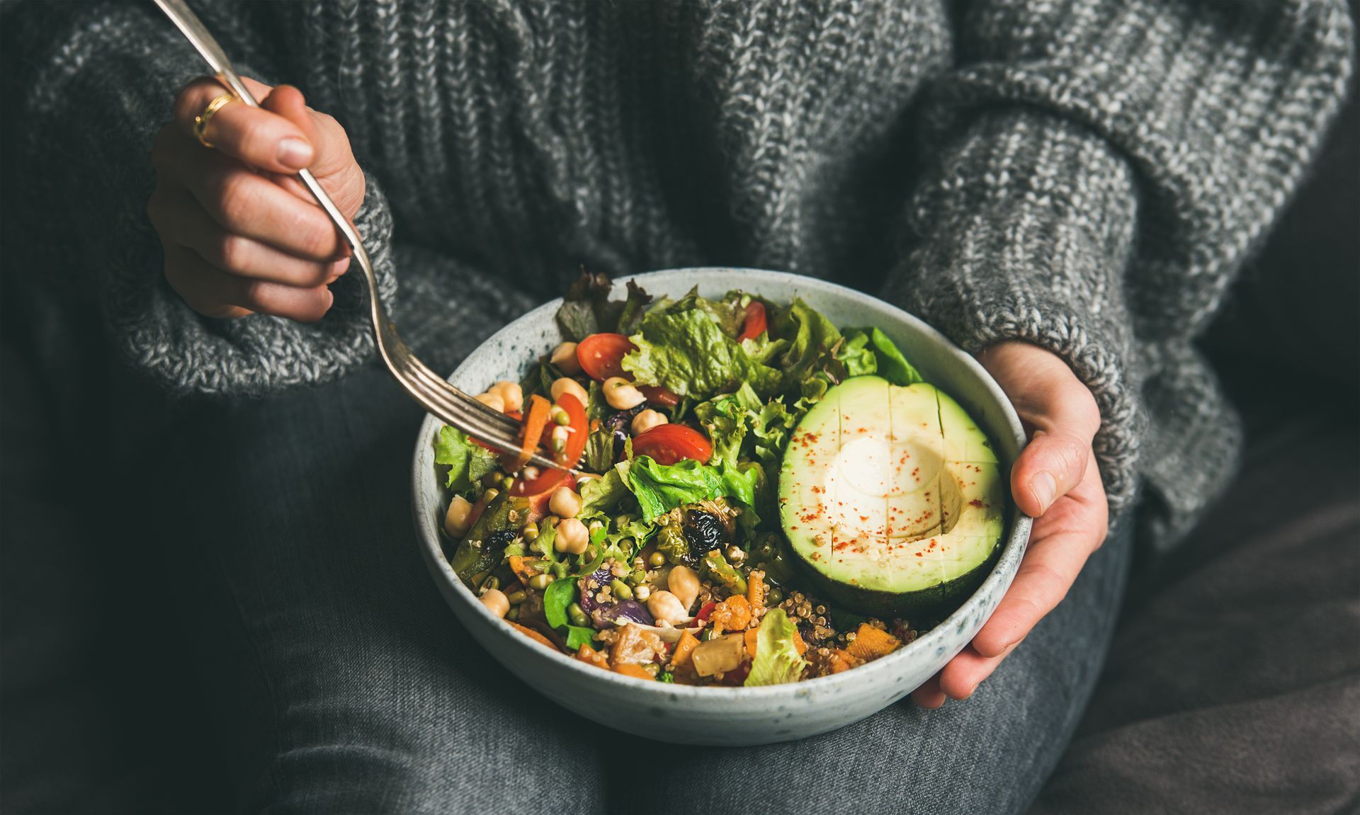 Person holding a bowl with a salad, avocado, and fork, in a gray sweater.