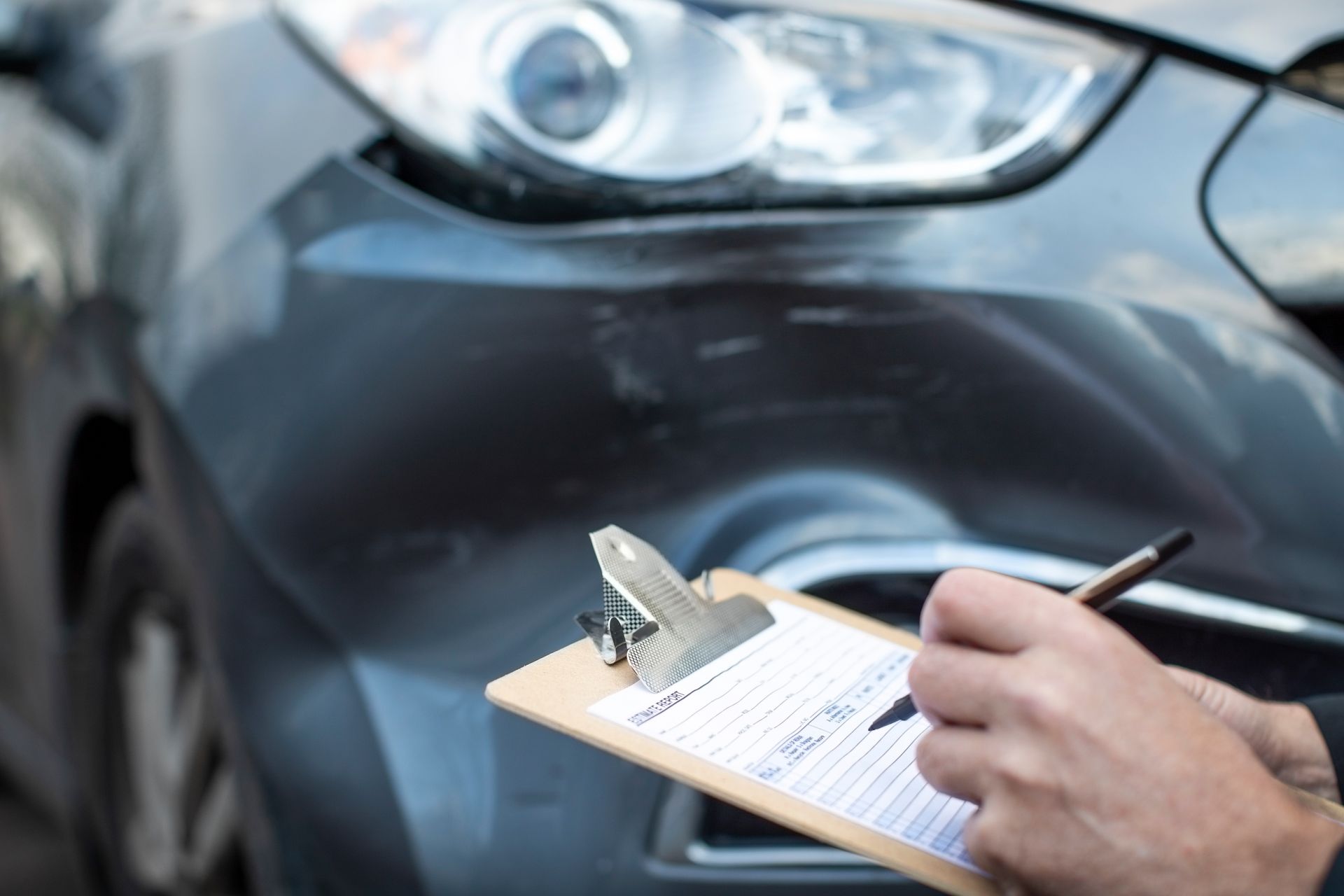 Person writing on a clipboard beside a gray car's front bumper and headlight
