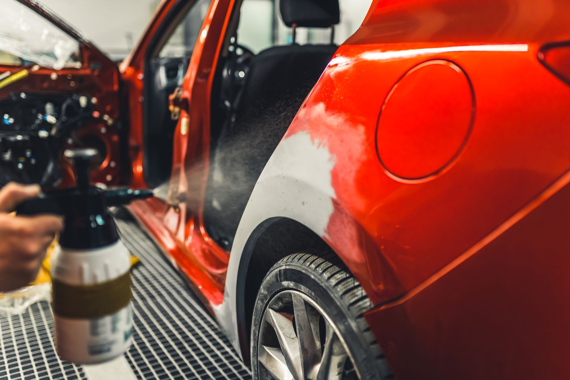 Person using a spray bottle to clean a red car’s rear fender in a garage