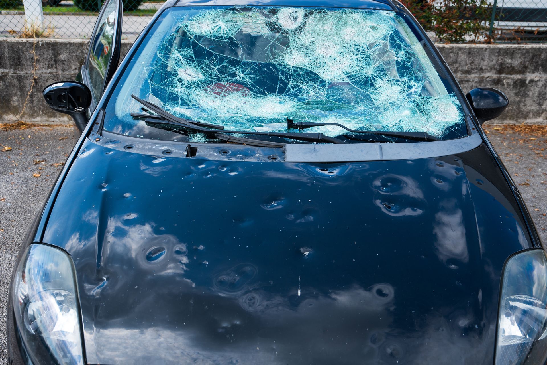 Black car with a shattered windshield and dented hood, covered in bullet holes, parked outdoors.