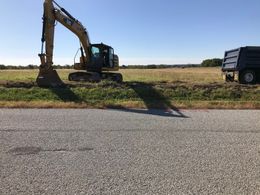 A yellow excavator sits in a grassy field next to a dark dump truck near a gravel road under a clear blue sky.