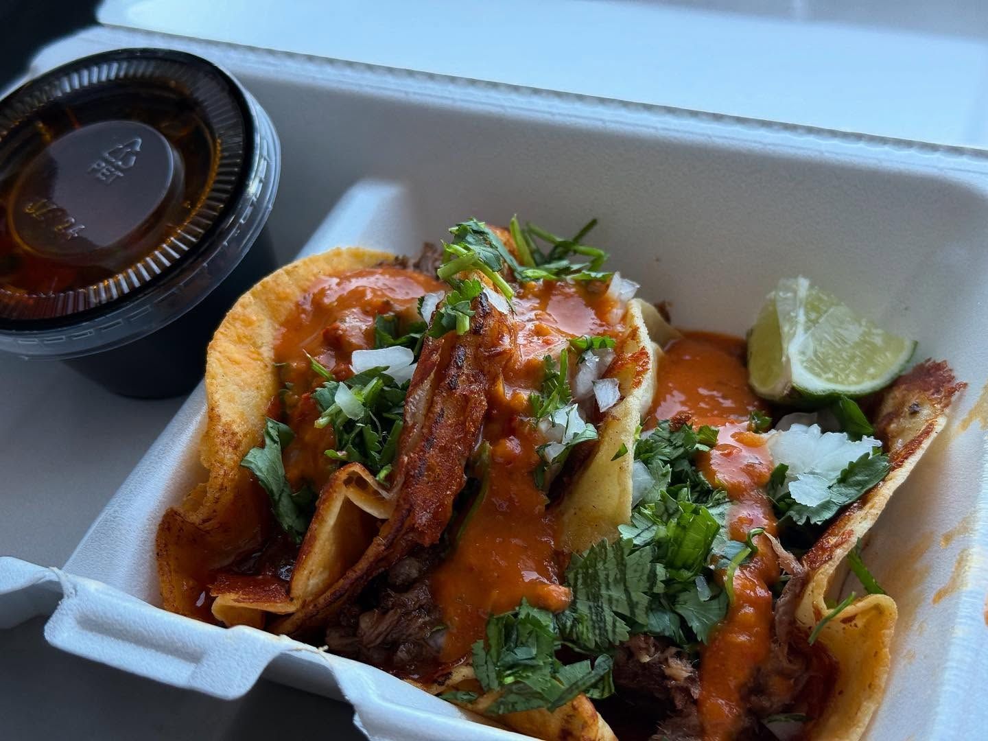 Three tacos in a white container, topped with red sauce, onions, cilantro, and a lime wedge. A side of sauce is visible.