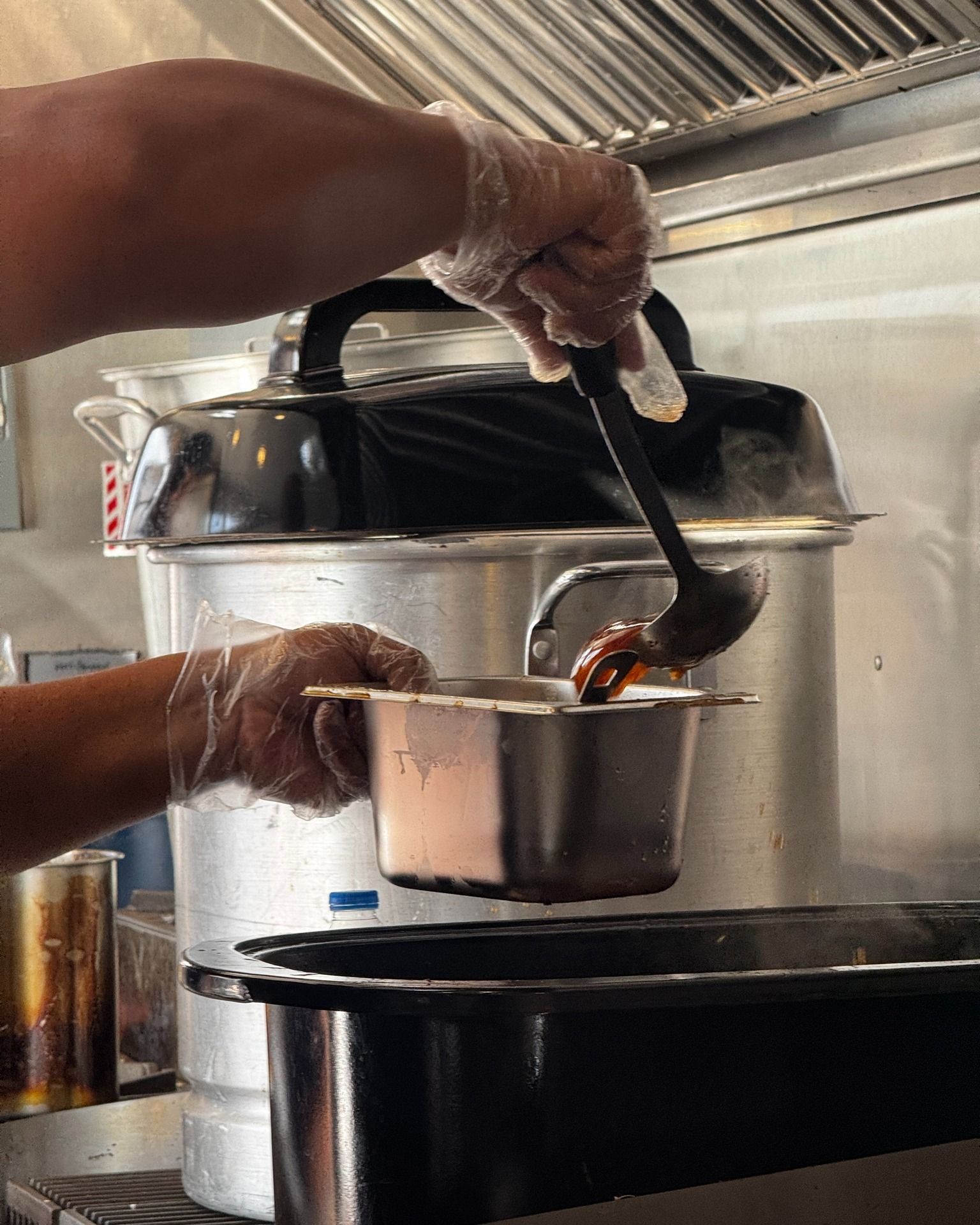 A person in gloves ladles food from a steam table into a metal container in a commercial kitchen.