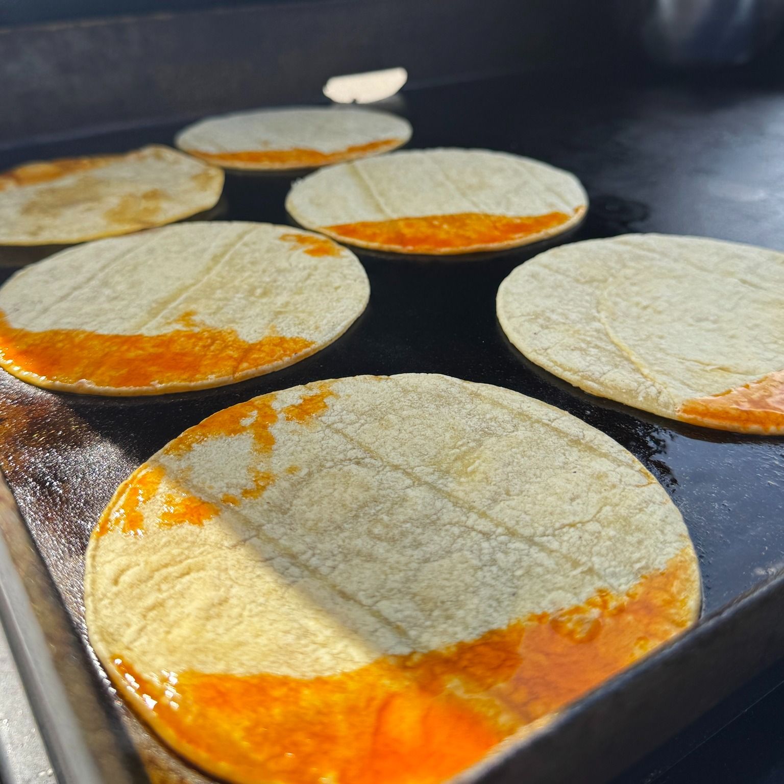 Corn tortillas on a hot griddle, partially coated in orange sauce.