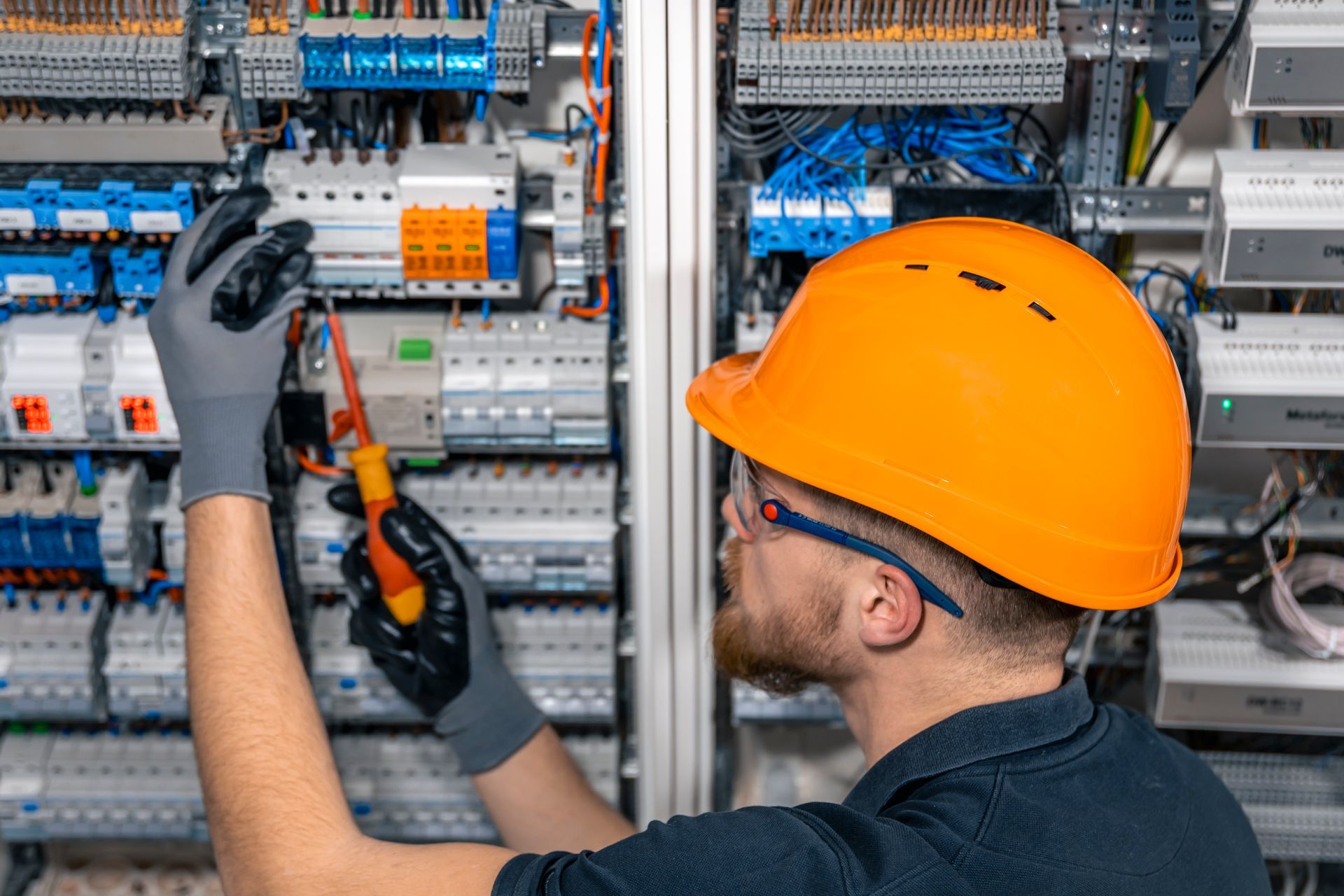Electrician in orange hard hat working on electrical panel, wearing gloves and safety glasses.