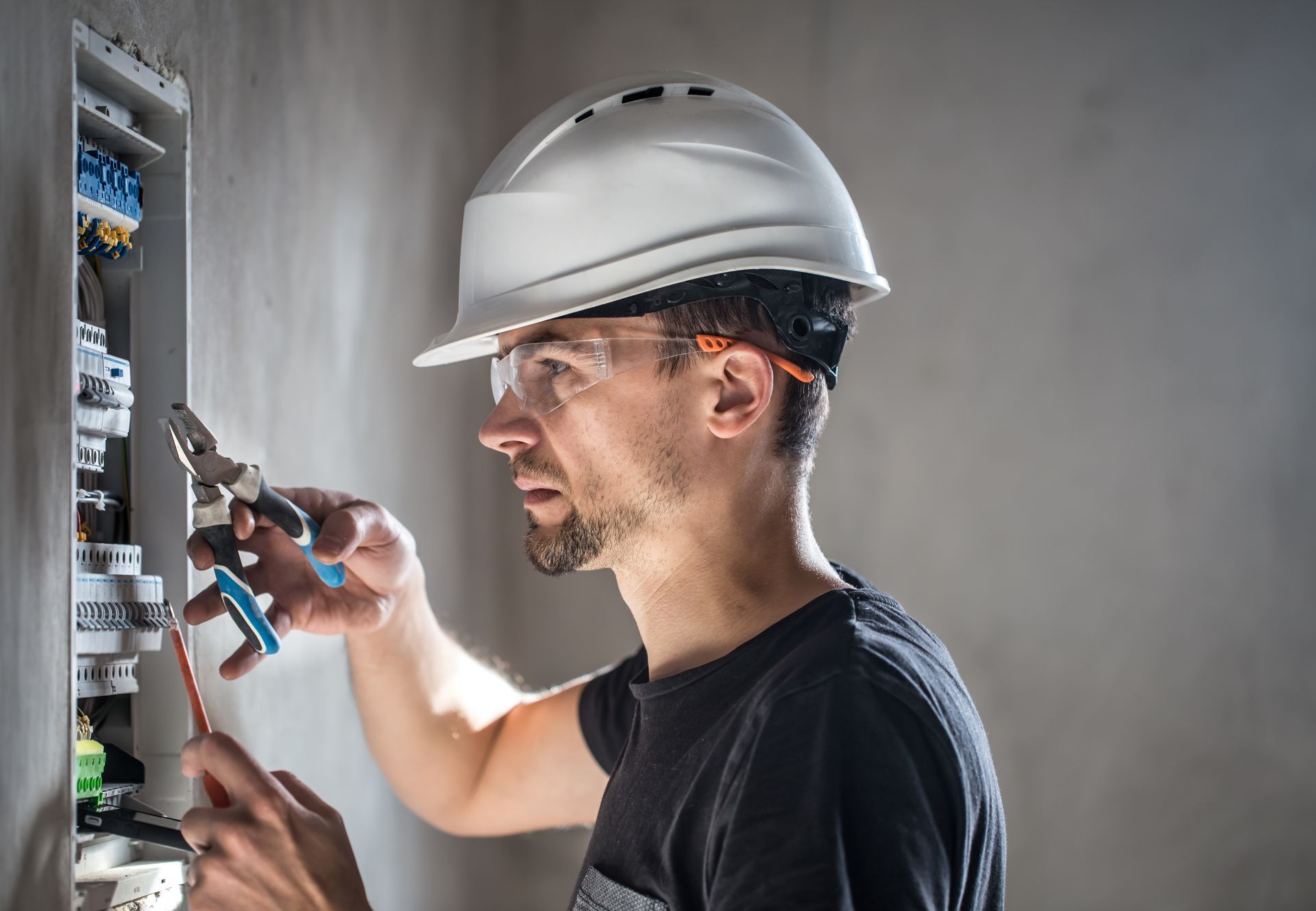 Electrician working on a circuit breaker panel, wearing a hard hat and safety glasses, indoor setting.