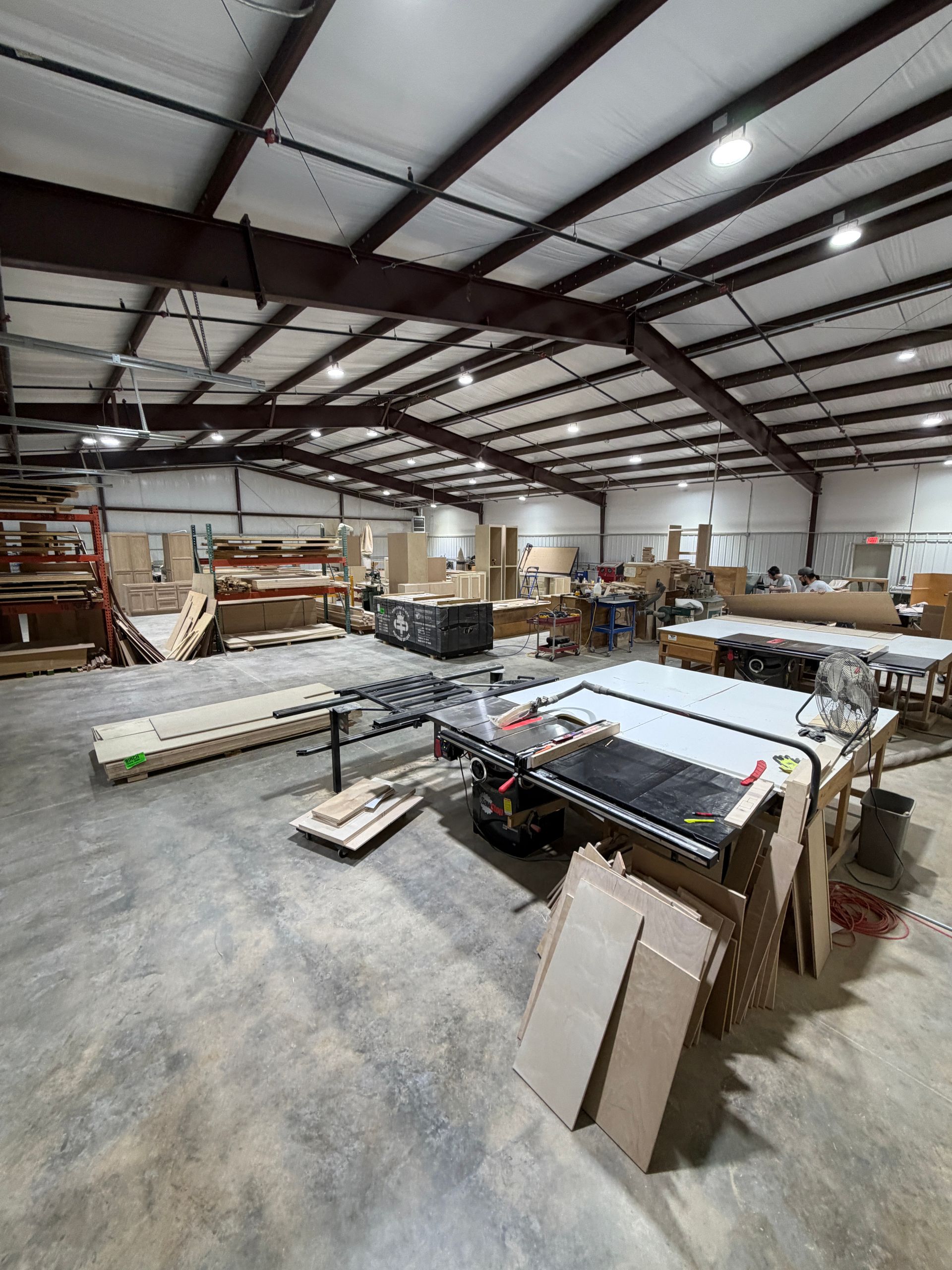 Large warehouse workshop with tables, tools, lumber, and materials under a metal beam ceiling.