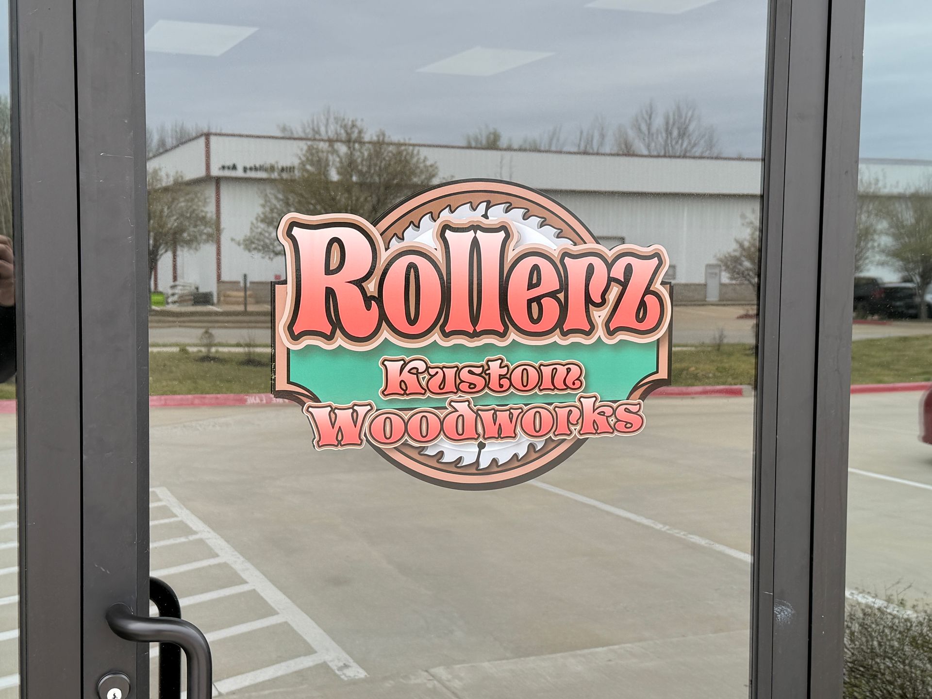 Rollerz Eastone Woodworks sign on a glass door, with a parking lot and building in the background