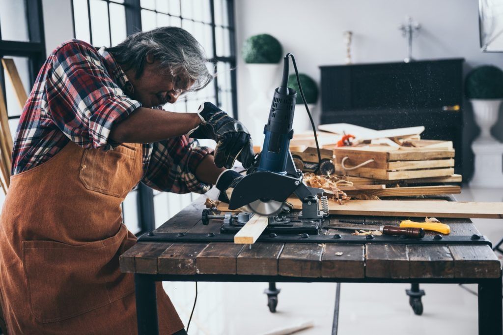 Person in apron leaning over a workbench, using a power tool in a workshop with scattered tools and materials.